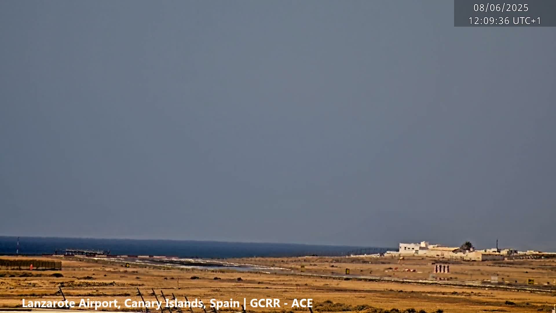 A flat, arid landscape with a distant ocean under a clear, light-grey sky shows a few low buildings near the coast.