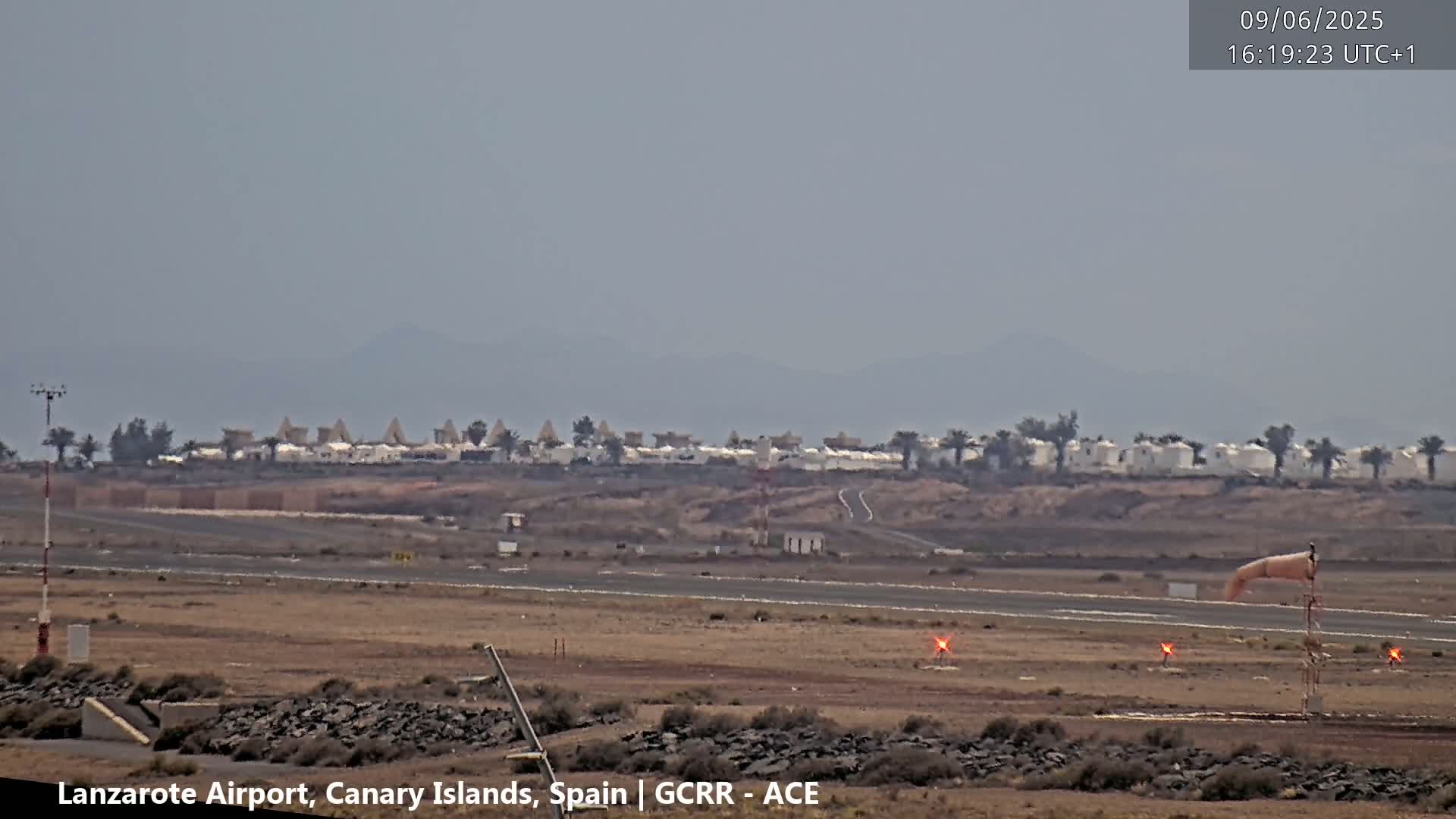 A hazy view of an airport runway with a windsock and distant buildings under a cloudy sky.