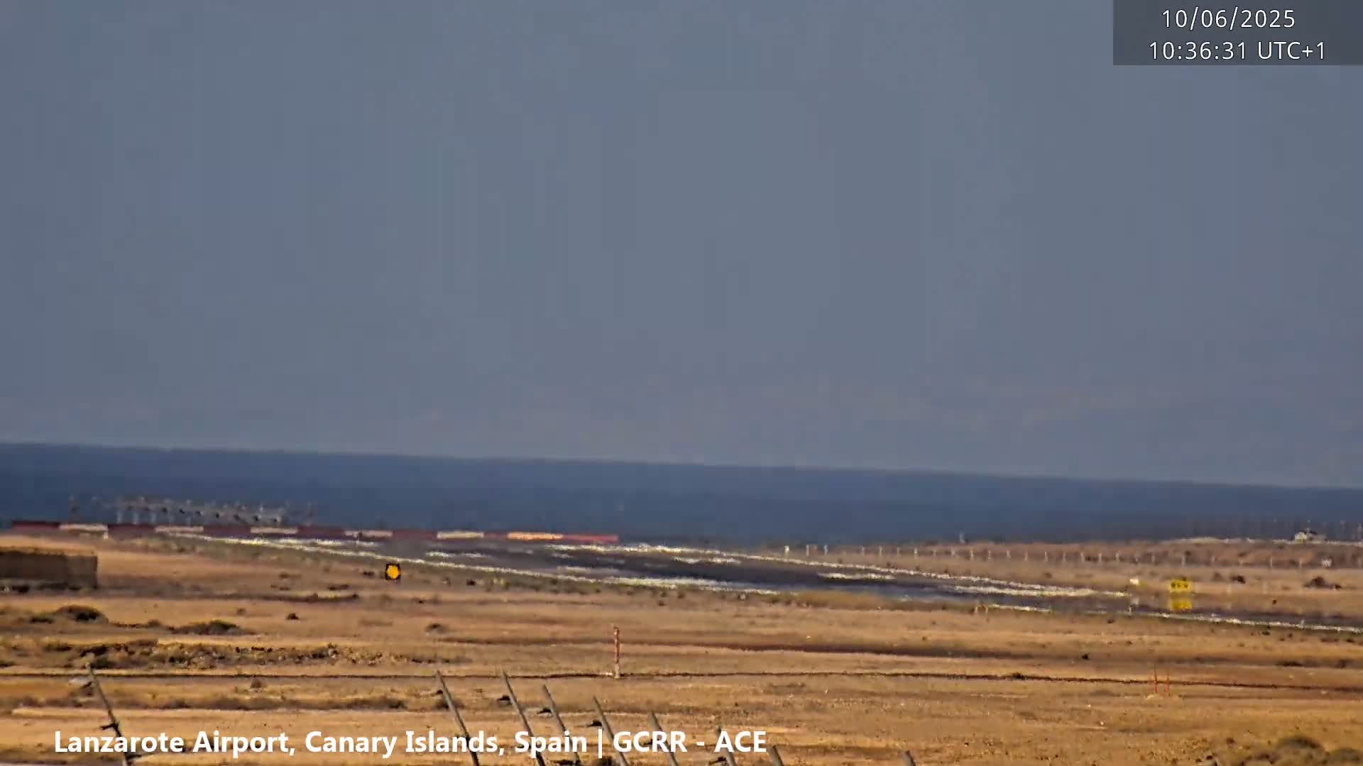 A flat, arid landscape with a calm ocean in the background under a clear, light-grey sky.