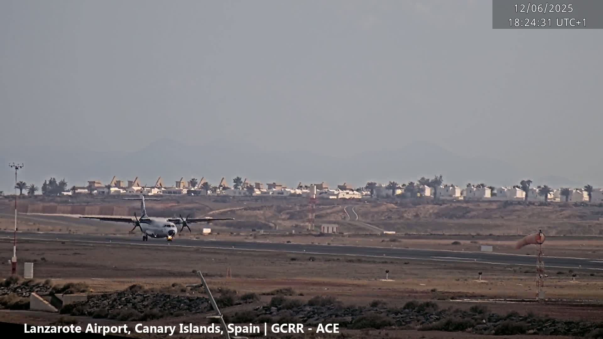 A turboprop airplane taxis on a runway, with hazy weather conditions and a line of low buildings visible in the background.