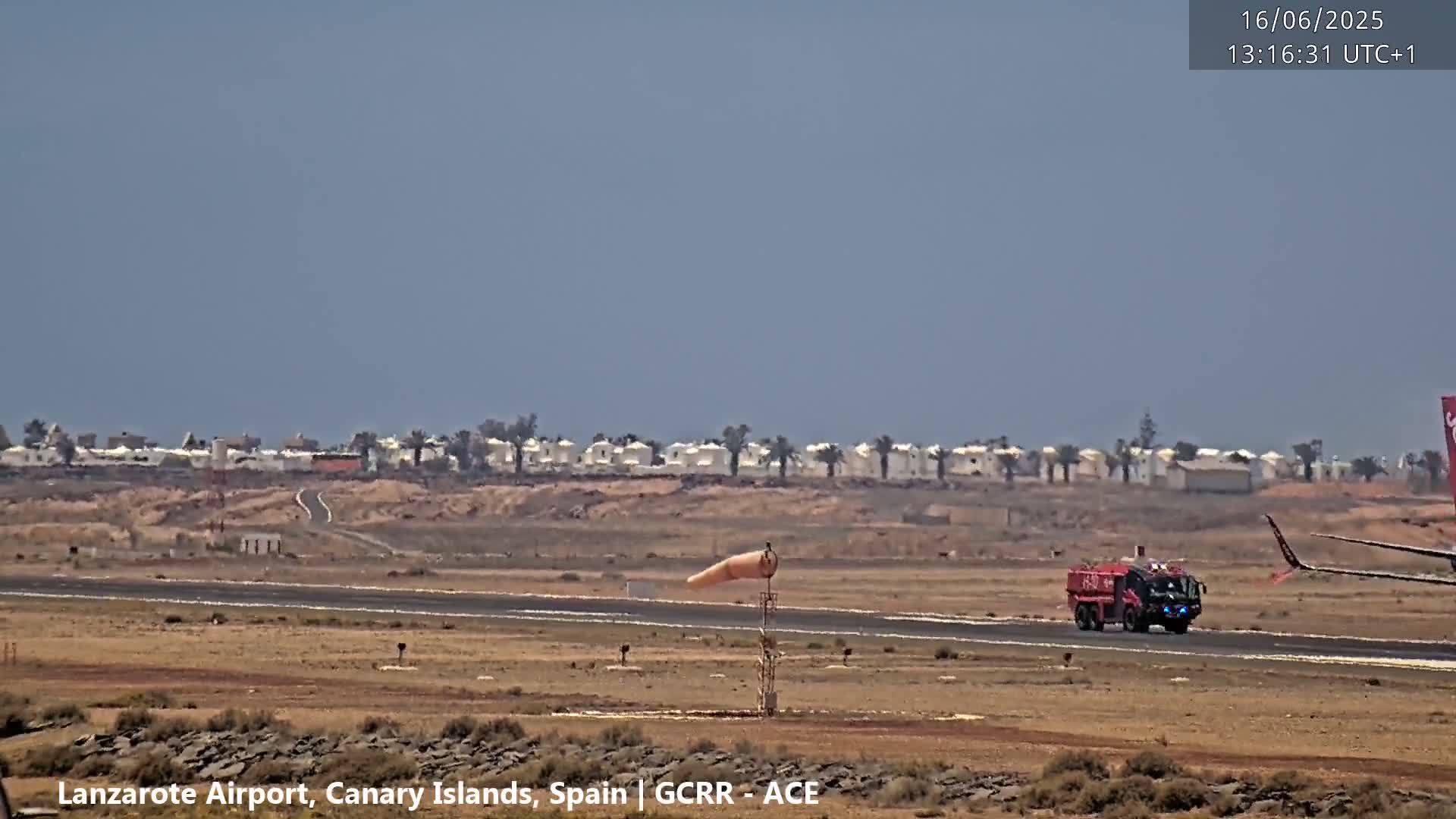 A red firetruck drives across a runway, a windsock indicates wind direction, and a row of white buildings are visible in the distance under a clear sky.