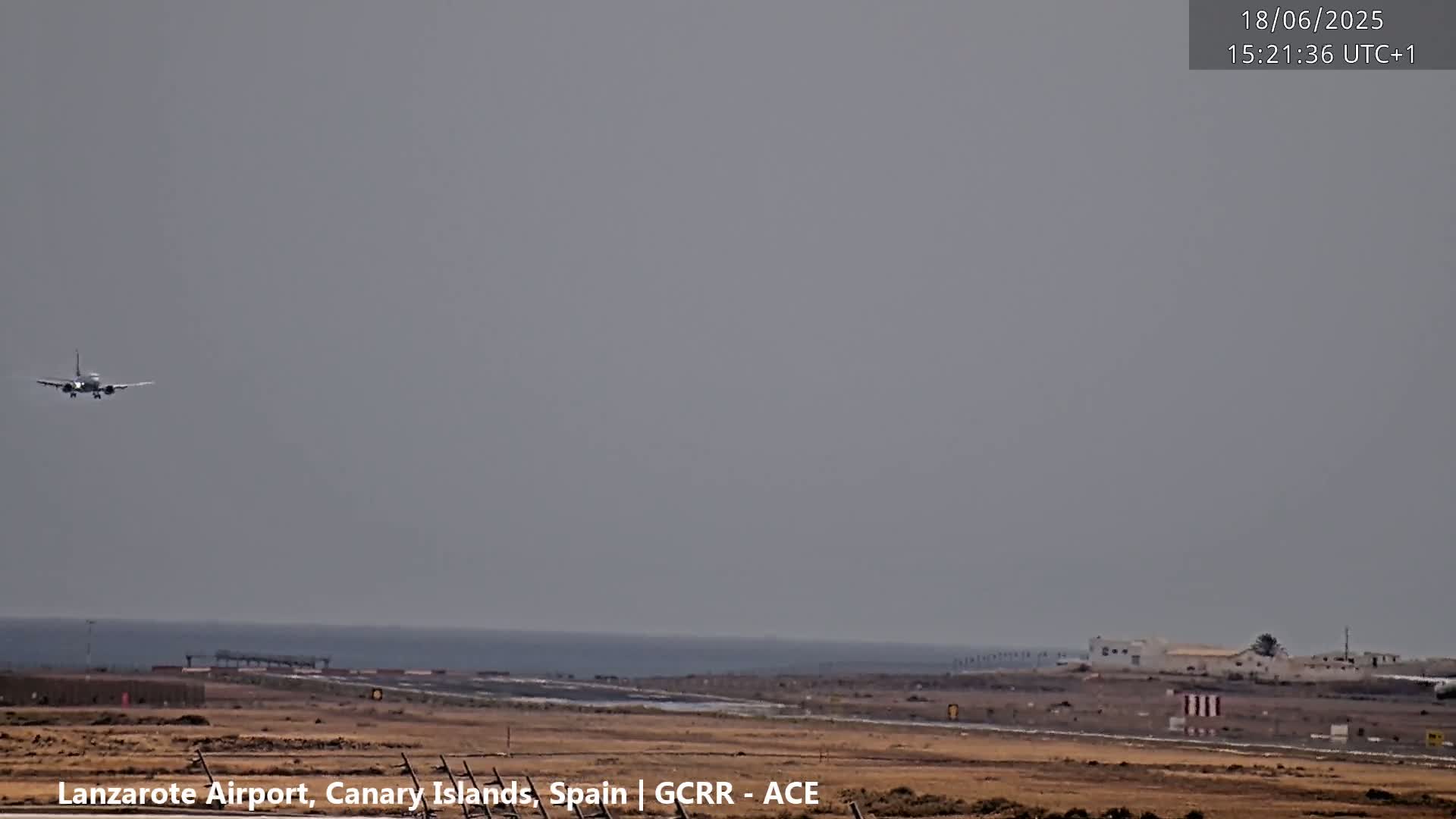 A passenger airplane is descending on a hazy day over a flat, arid landscape near a coastline.