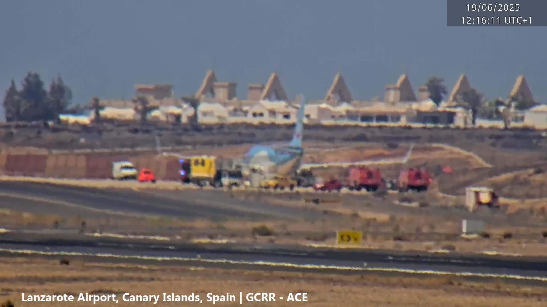 A damaged airplane is surrounded by emergency vehicles on an airport tarmac under a hazy sky.