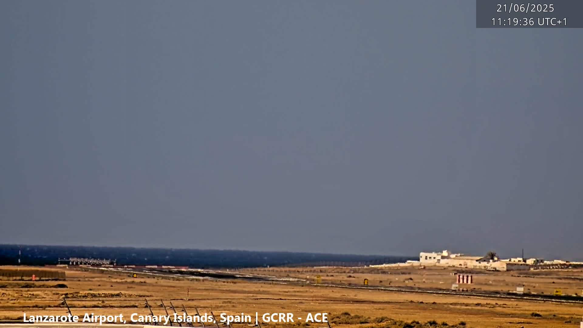A flat, arid landscape with a distant ocean under a hazy, light-grey sky shows a runway and airport buildings.