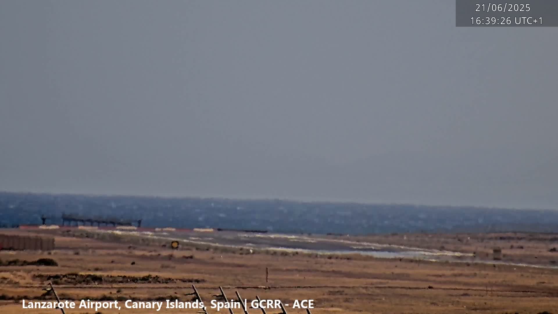 A hazy, distant view of a calm ocean and flat, brown land with a section of airport runway in the foreground.