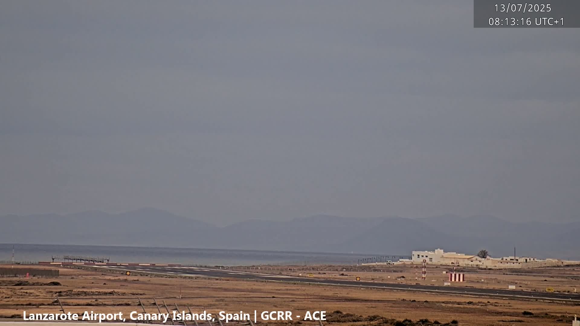 A hazy view of an airport runway extends to a calm ocean and distant mountains under a gray sky.