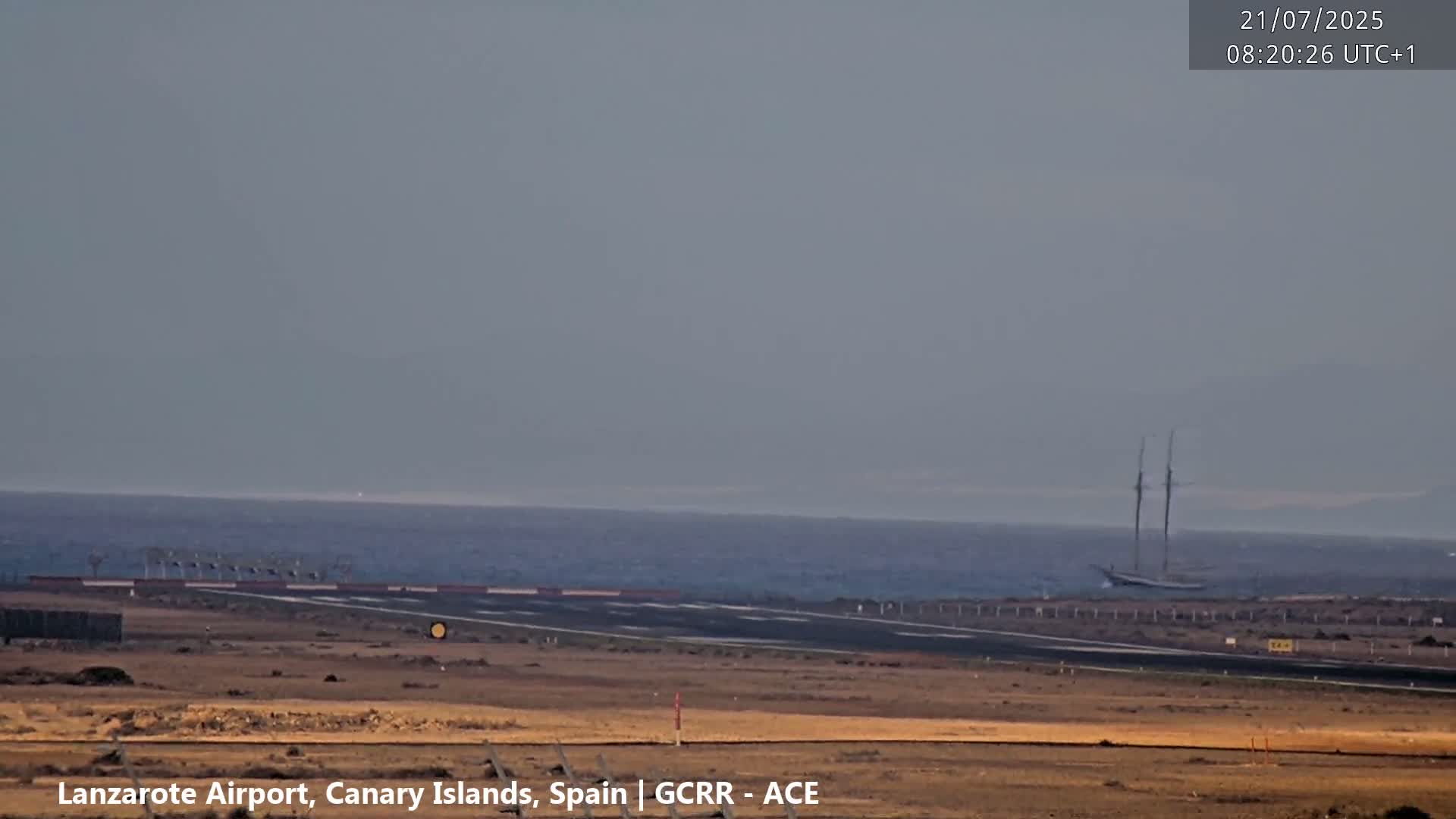 A large sailboat is visible on the ocean in the distance from an airport runway on a hazy day.