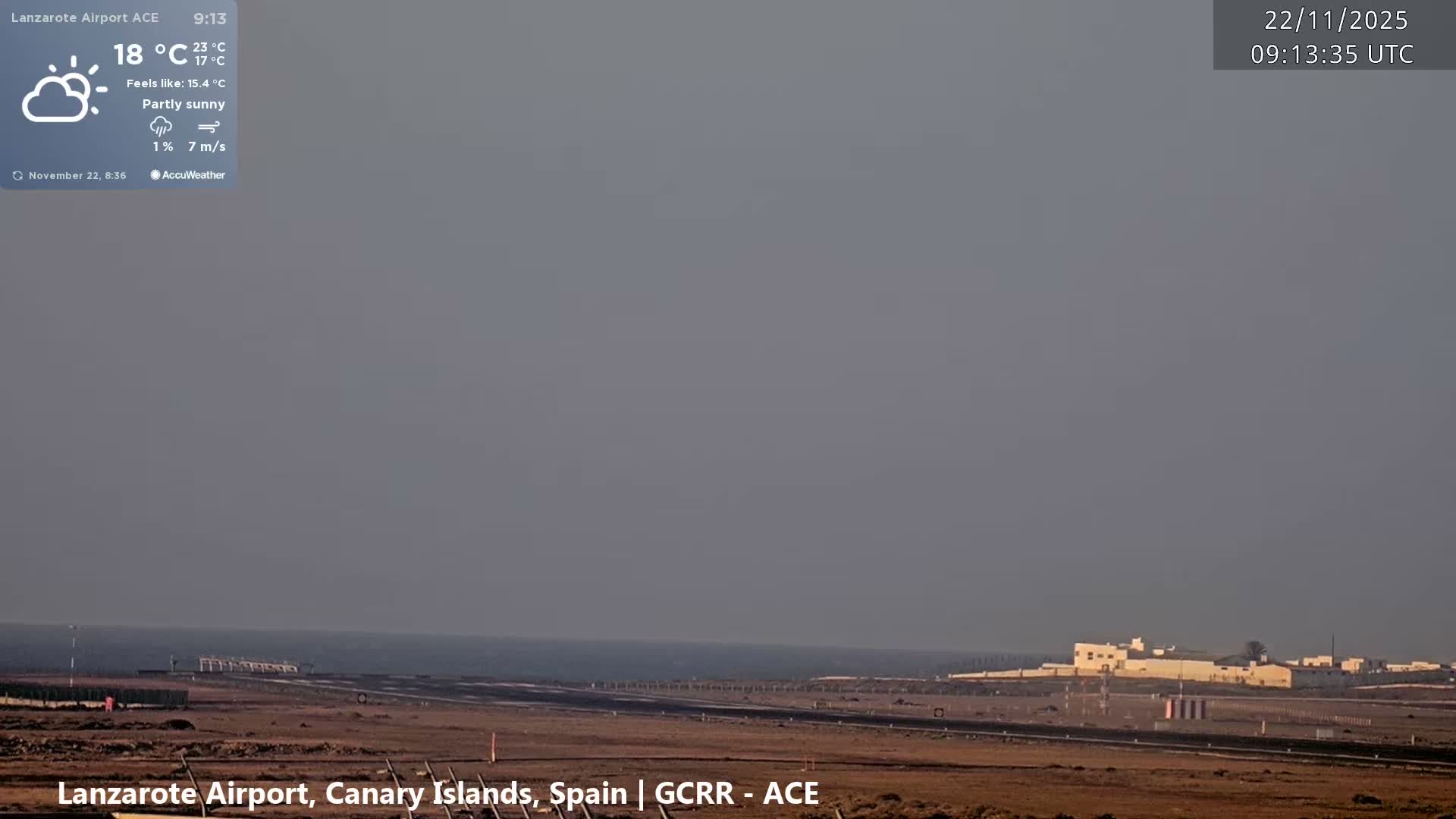 A wide, outdoor view captures an airport runway stretching towards the calm sea under a hazy gray sky, with a few white buildings visible on the distant right horizon.