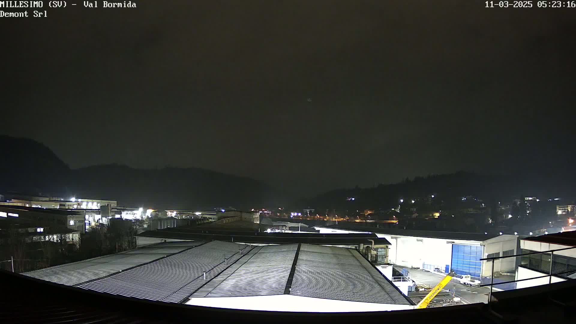 A nighttime view of industrial buildings and a hillside under a dark, overcast sky.
