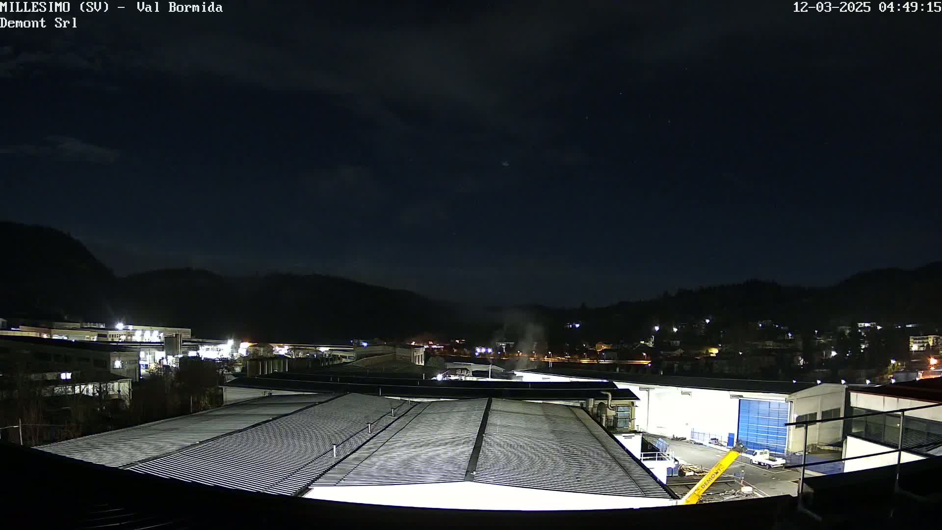 A nighttime view of an industrial area with buildings and hills in the background under a clear, dark sky.