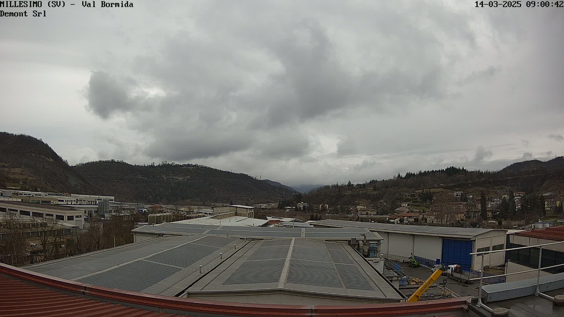 A view from a rooftop shows overcast skies over a valley with industrial buildings and a small town nestled between hills.