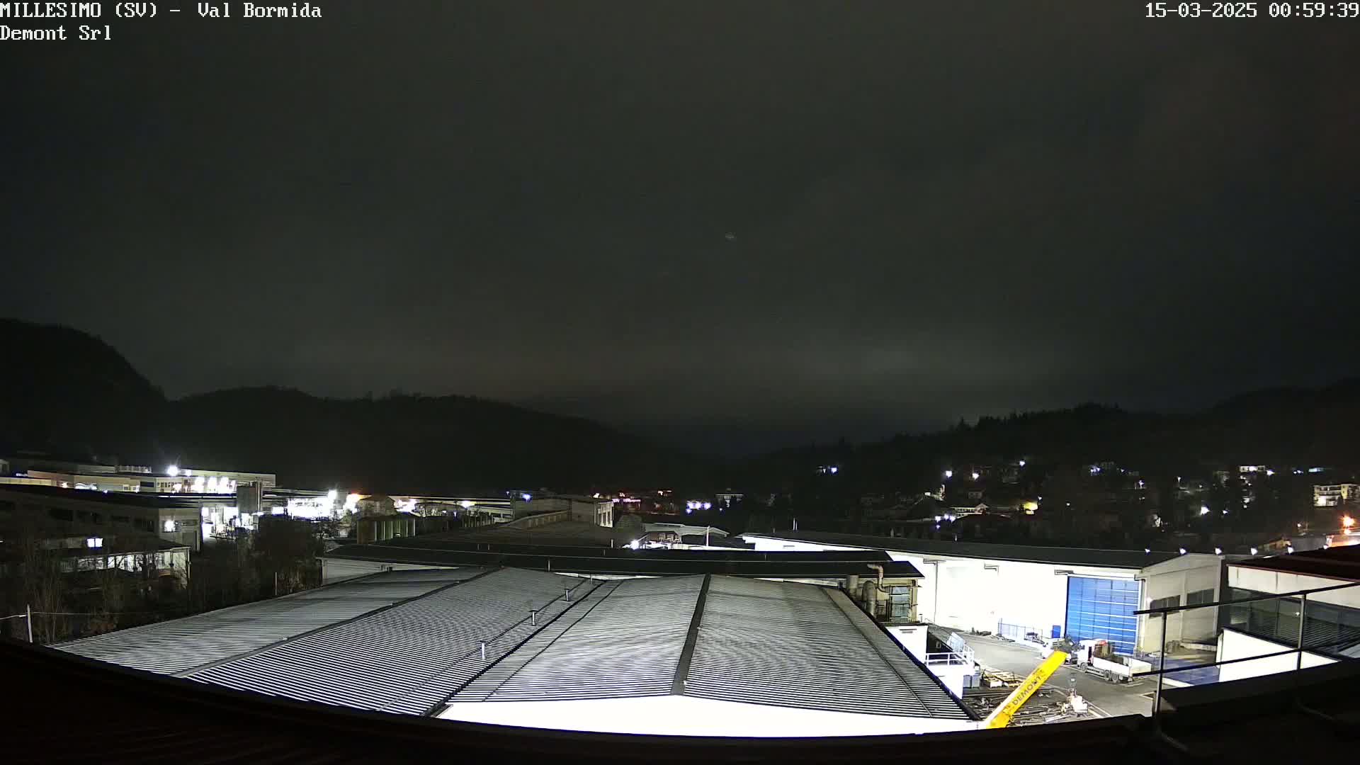A nighttime view of an industrial area nestled between dark hills under a mostly cloudy sky.