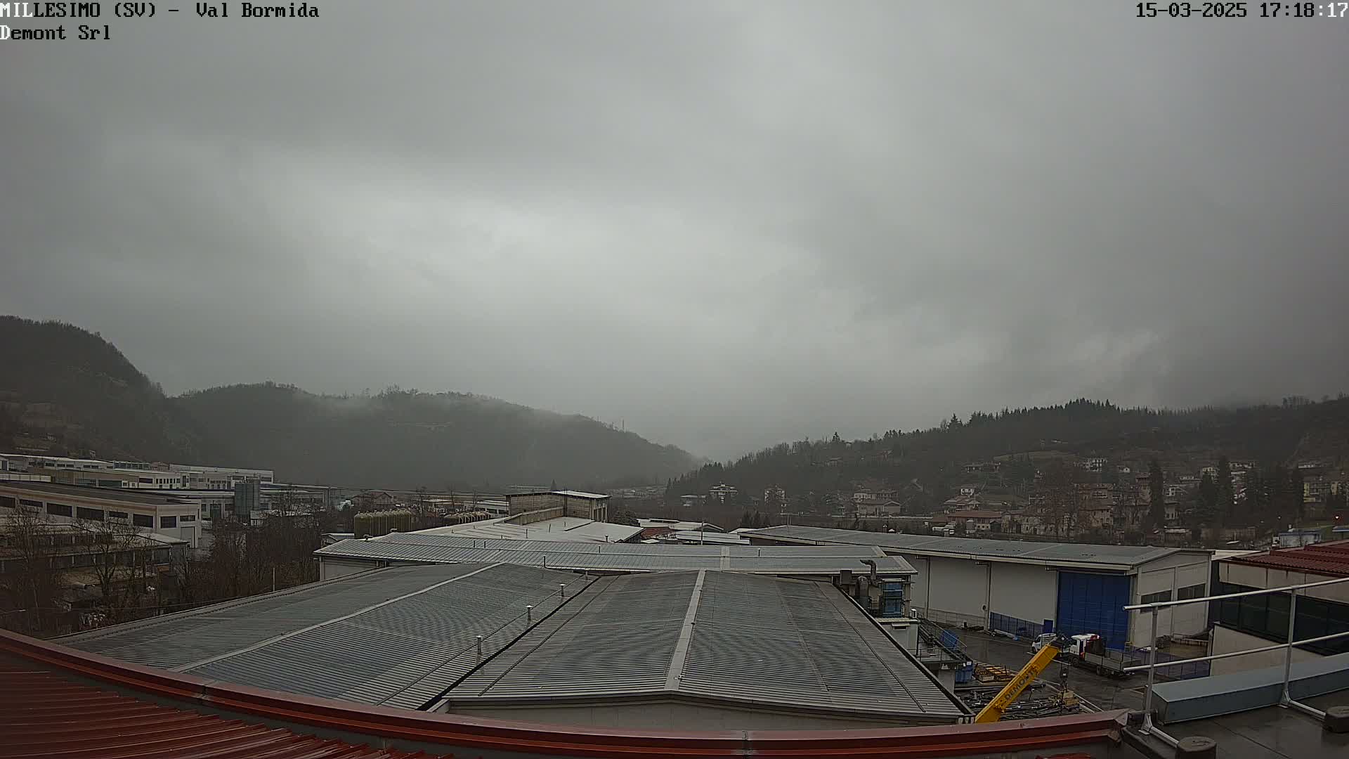 A view from a rooftop shows overcast skies over a valley of industrial buildings and residential houses nestled at the base of misty mountains.