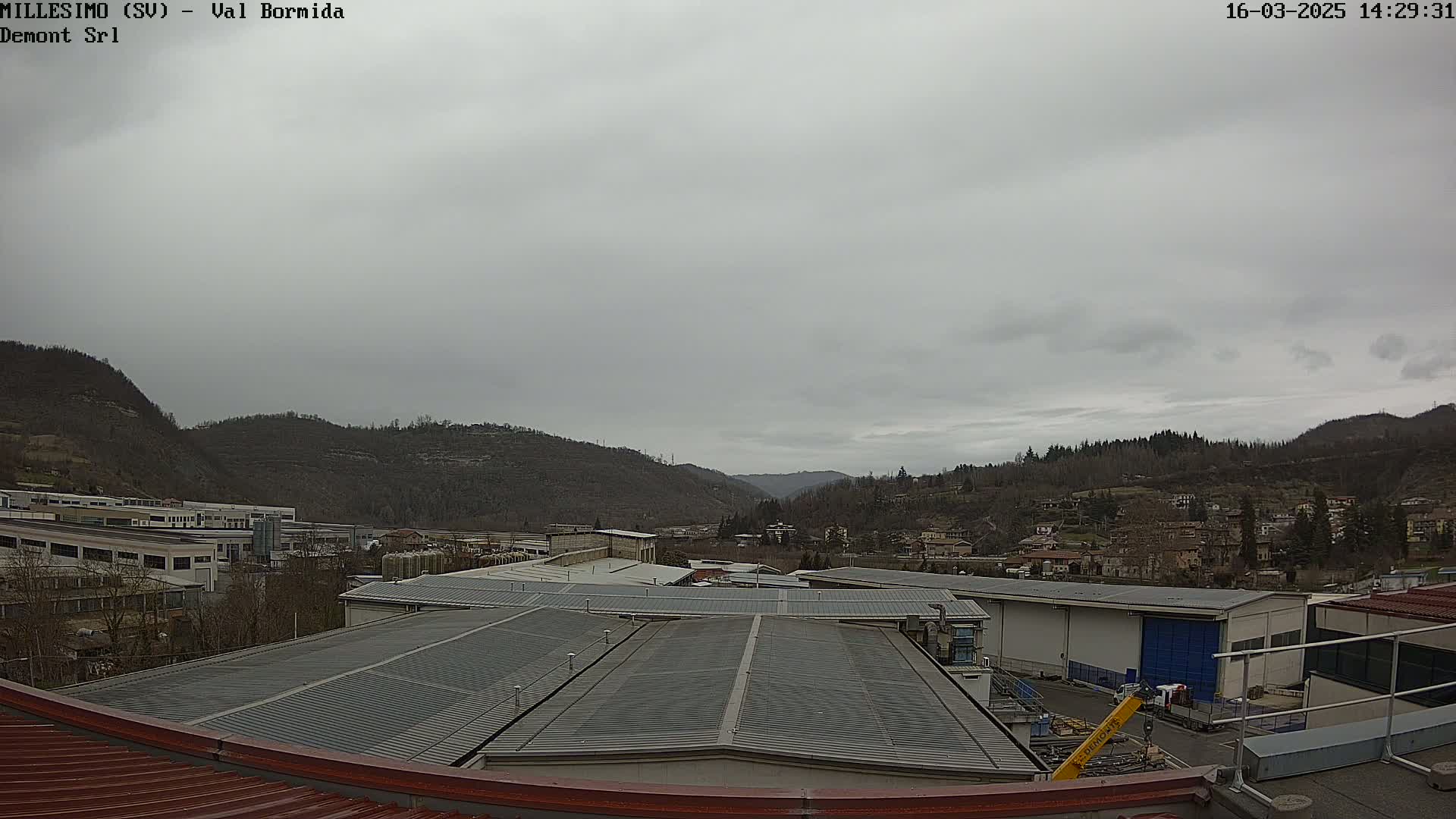 A view from a rooftop shows a cloudy sky over industrial buildings and a town nestled in a valley between hills.