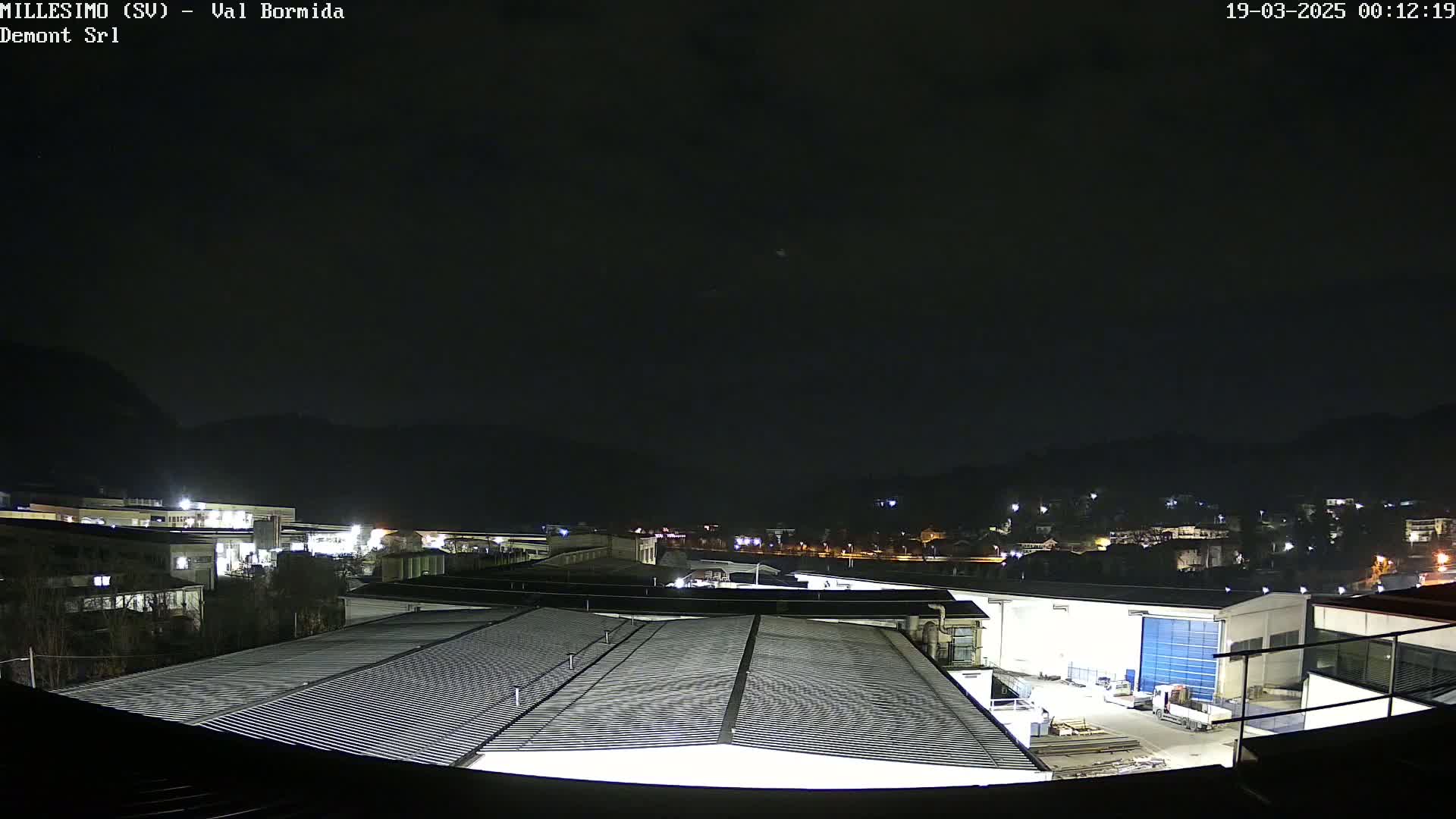 A nighttime view of industrial buildings and a town nestled against dark hills under a clear, dark sky.
