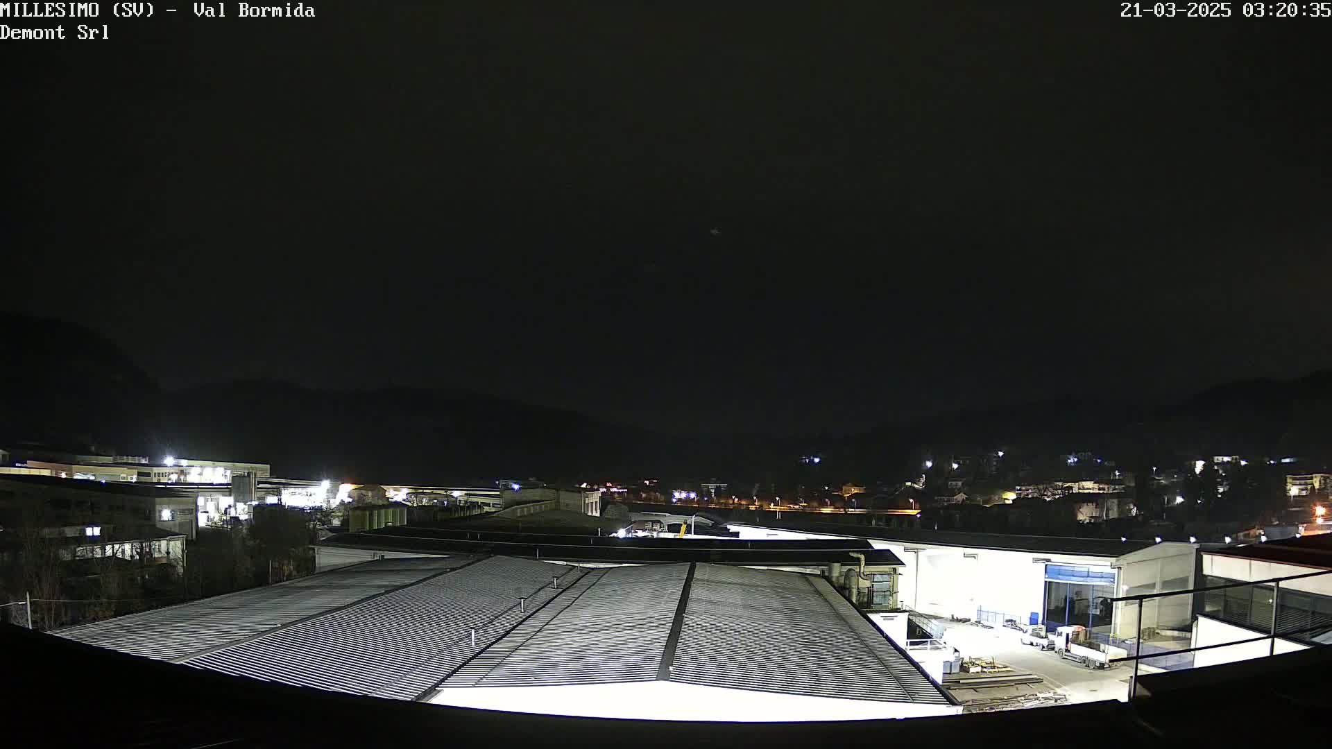 A nighttime view of an industrial area with buildings and a corrugated metal roof in the foreground, under a dark, clear sky.
