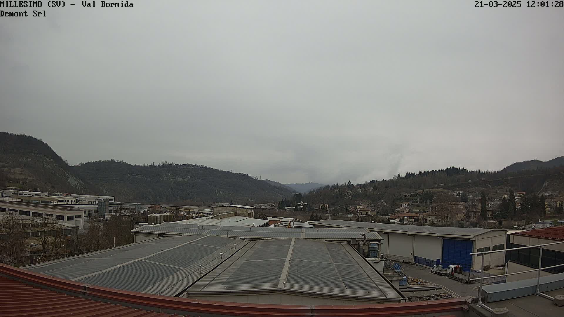 A view from a rooftop shows industrial buildings and a town nestled in a valley under an overcast sky.