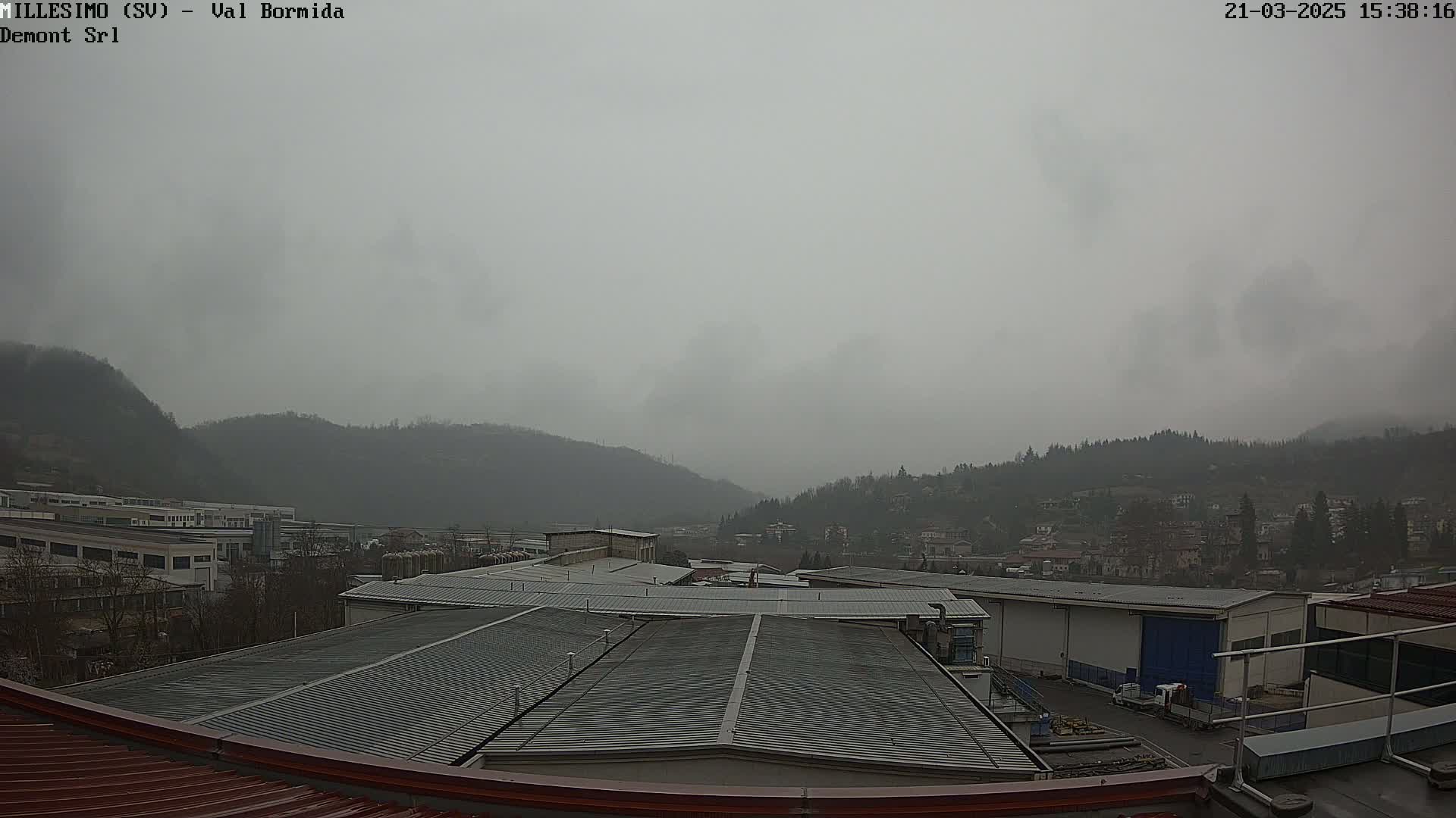 A view of industrial buildings and a town nestled in a valley under a heavily overcast sky.