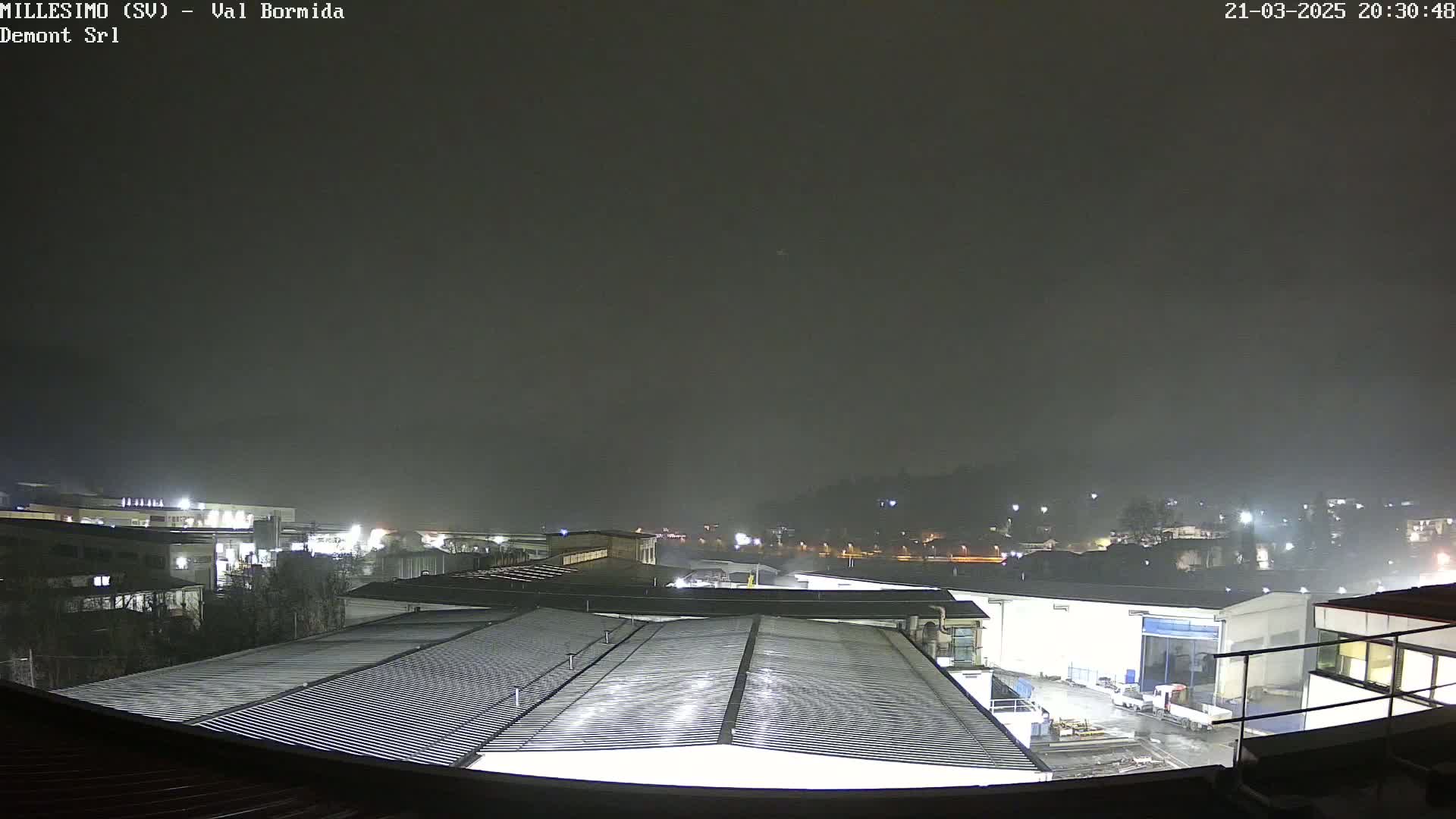 A nighttime view of an industrial area under a dark, possibly foggy or misty sky, showing several buildings with illuminated rooftops and surrounding grounds.
