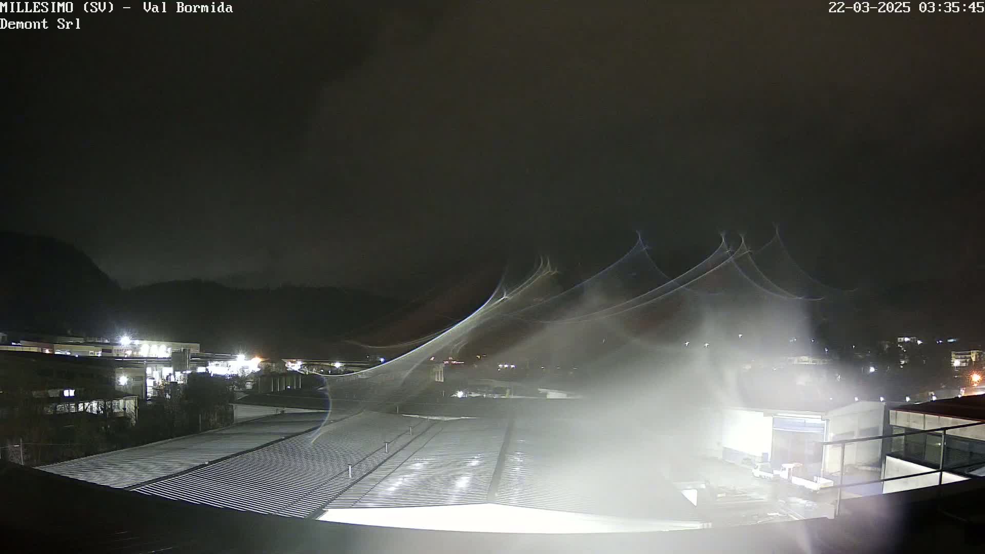 A nighttime view of an industrial area with a corrugated metal roof in the foreground, under a dark, misty sky with streaks of light possibly from rain or snow.