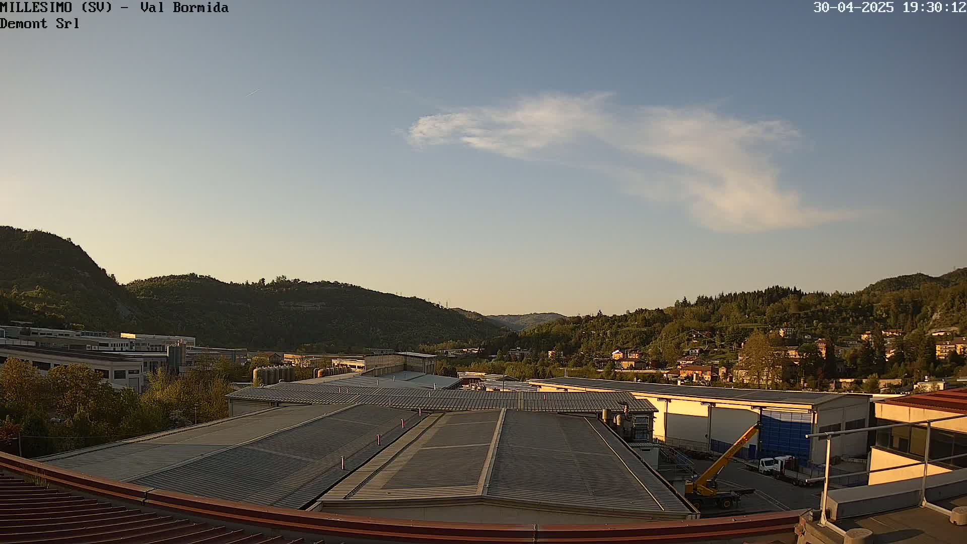 A mostly clear evening sky over a valley of green hills and industrial buildings with a few scattered clouds.
