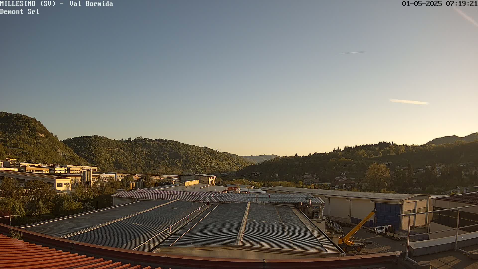 A clear, early evening sky overlooks a valley filled with green hills and industrial buildings, with a yellow crane visible near some structures.