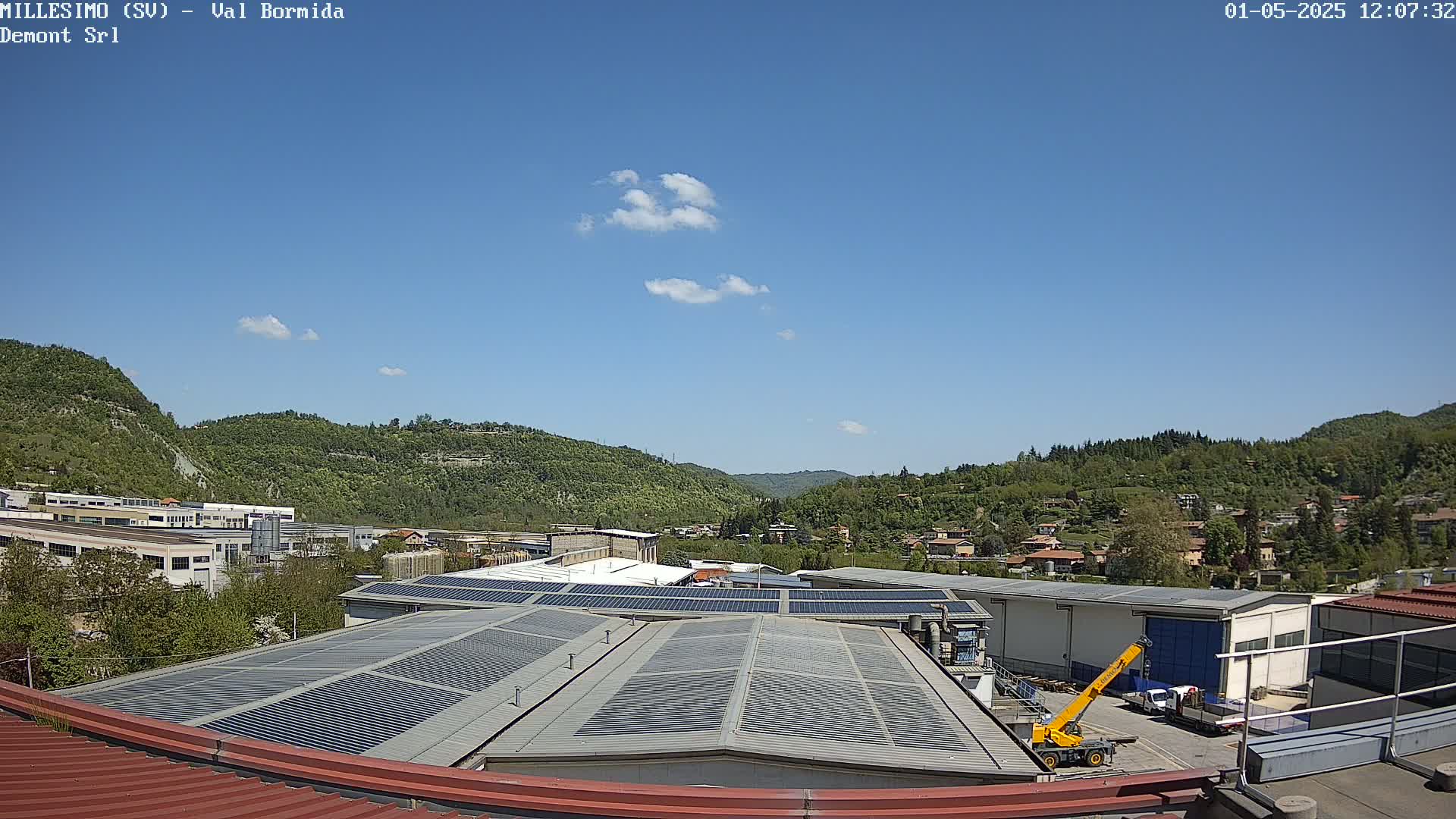 A mostly sunny day reveals a view of a factory with solar panels on its roof, nestled in a valley surrounded by green hills and a small village.
