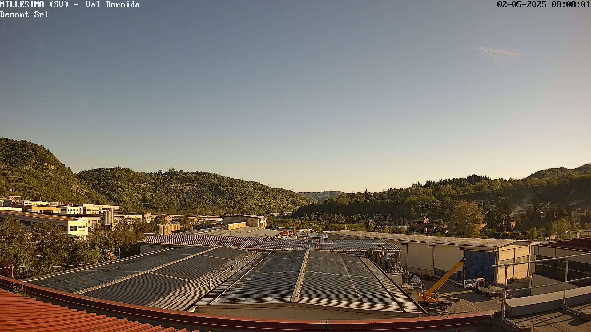 A clear, sunny day reveals a valley of green hills and industrial buildings with solar panels on rooftops.
