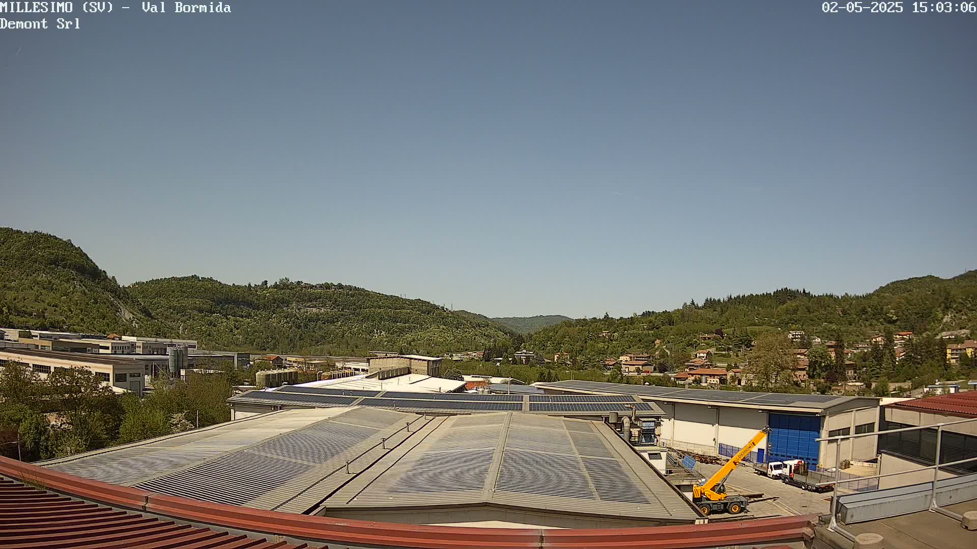 A clear, sunny day reveals an industrial area with buildings featuring solar panels on their roofs, nestled in a valley surrounded by green hills.