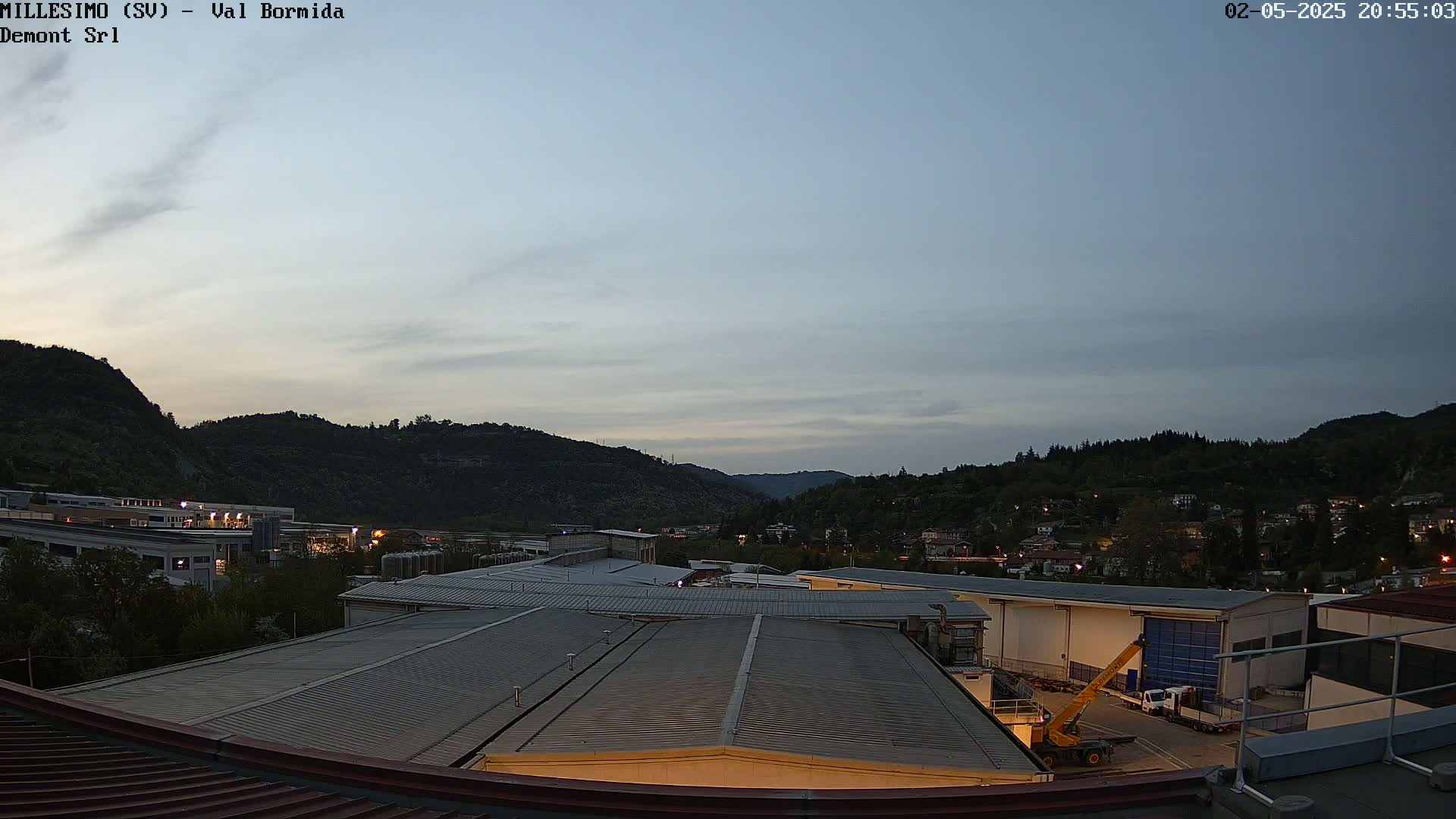 A twilight view shows a flat, gray industrial rooftop overlooking a valley of dark green hills and a town with scattered lights under a pale sky.
