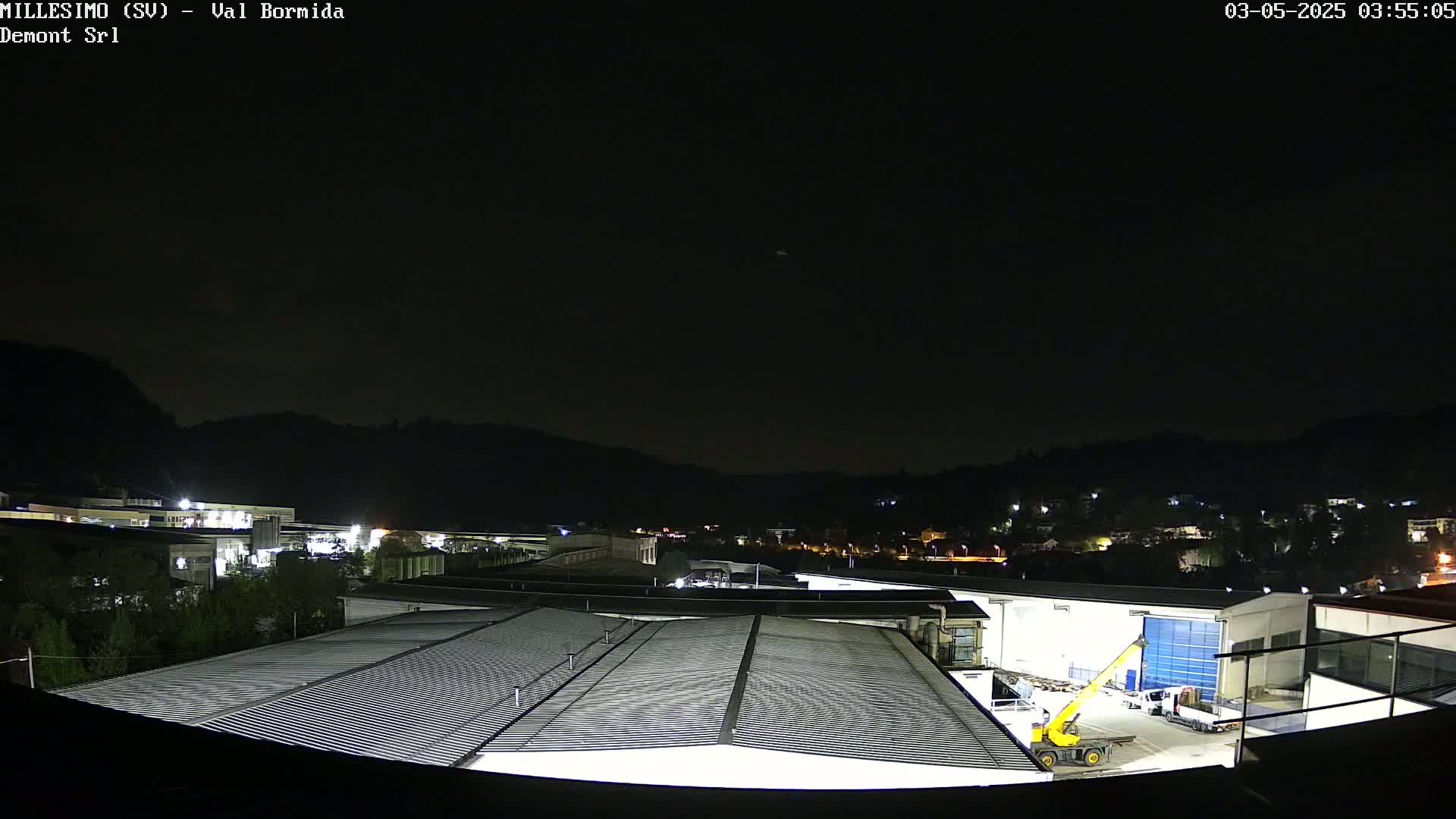 A nighttime view of an industrial area with several buildings, a yellow crane, and some vehicles, all under a dark, clear sky.