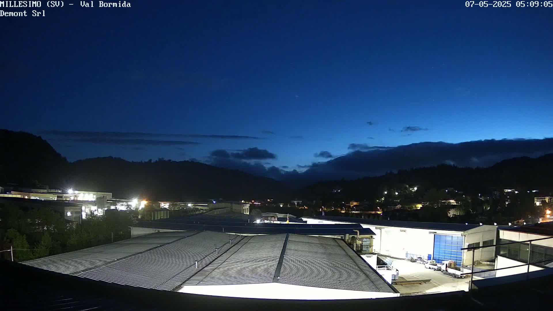 A nighttime view of an industrial area with a corrugated metal roof in the foreground, under a dark blue, mostly clear sky with some clouds near the horizon, and mountains in the background.