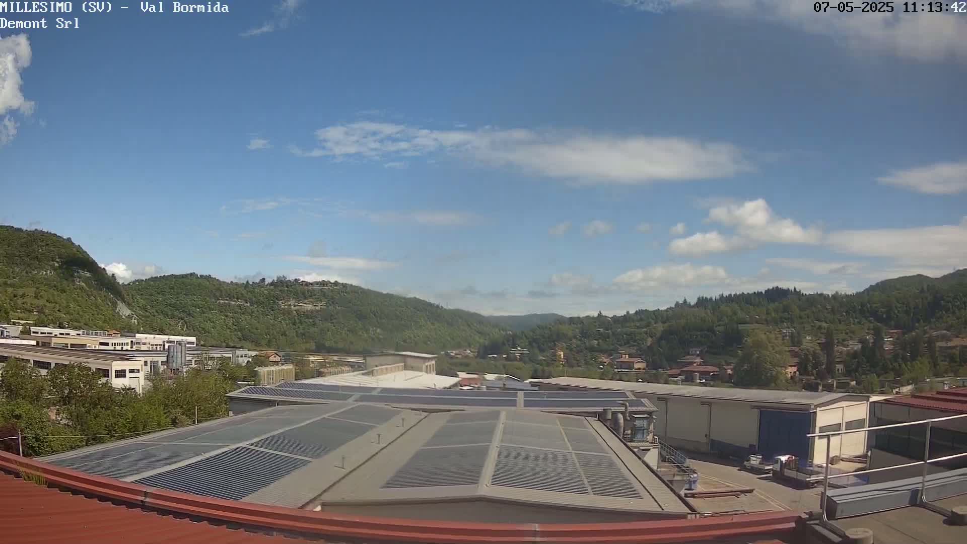 A mostly sunny day shows a view of industrial buildings and a valley filled with green hills and a few houses under a partly cloudy sky.