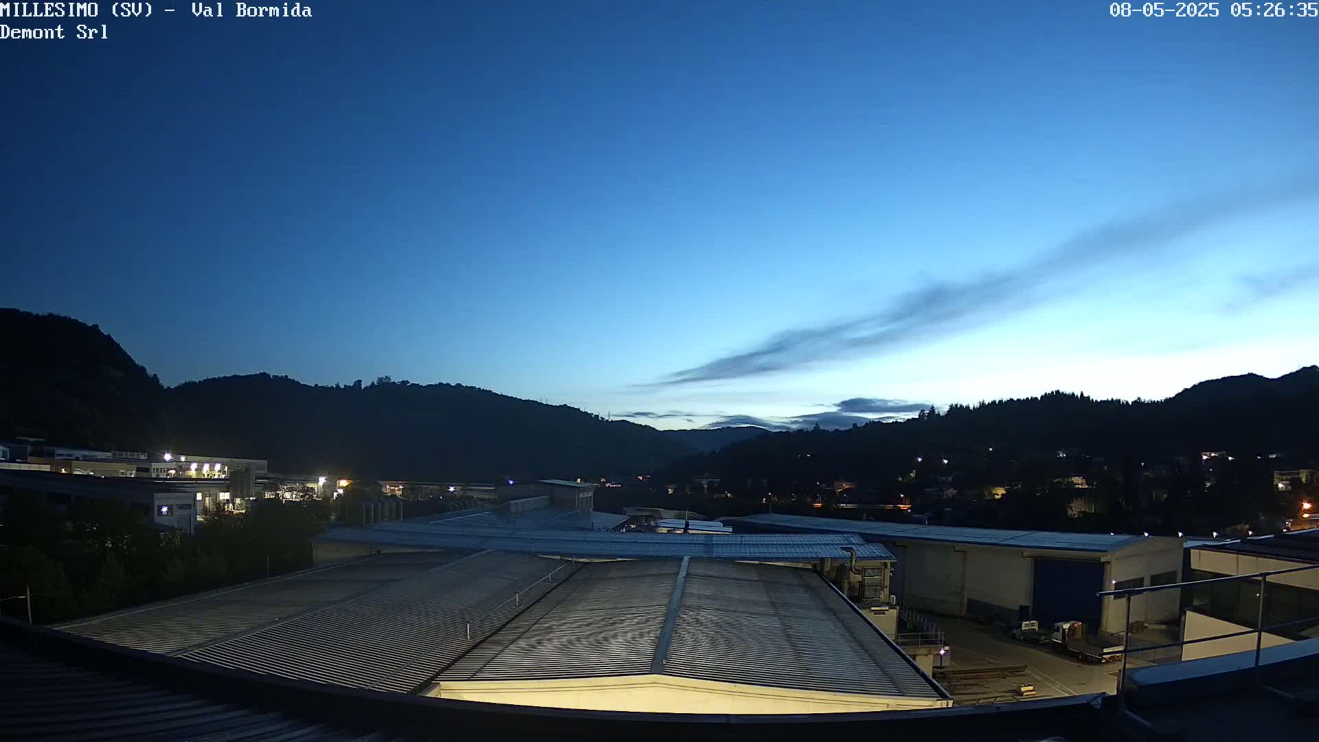 A twilight view shows a sprawling industrial area nestled against dark, tree-covered hills under a mostly clear, deep blue sky.