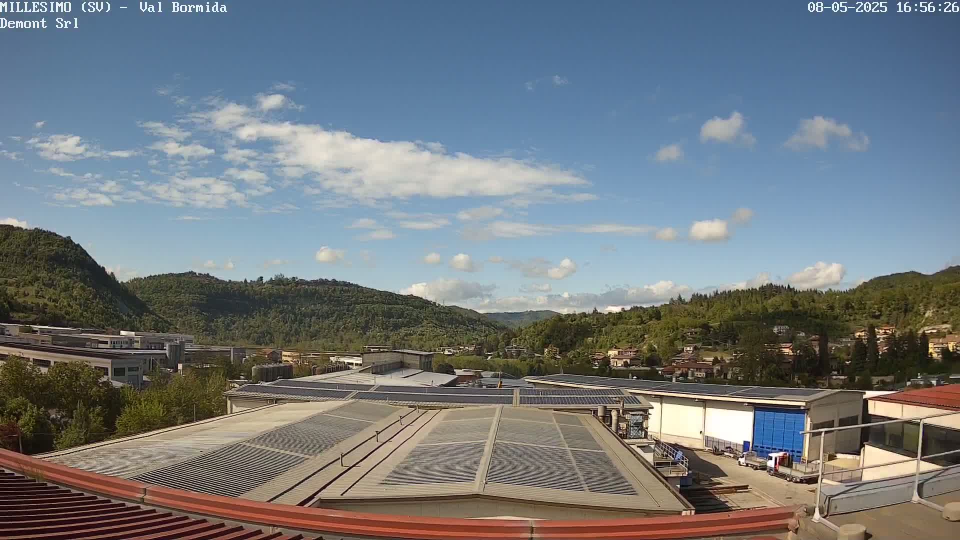A view from a rooftop shows industrial buildings with solar panels, surrounded by green hills under a mostly sunny sky with scattered clouds.