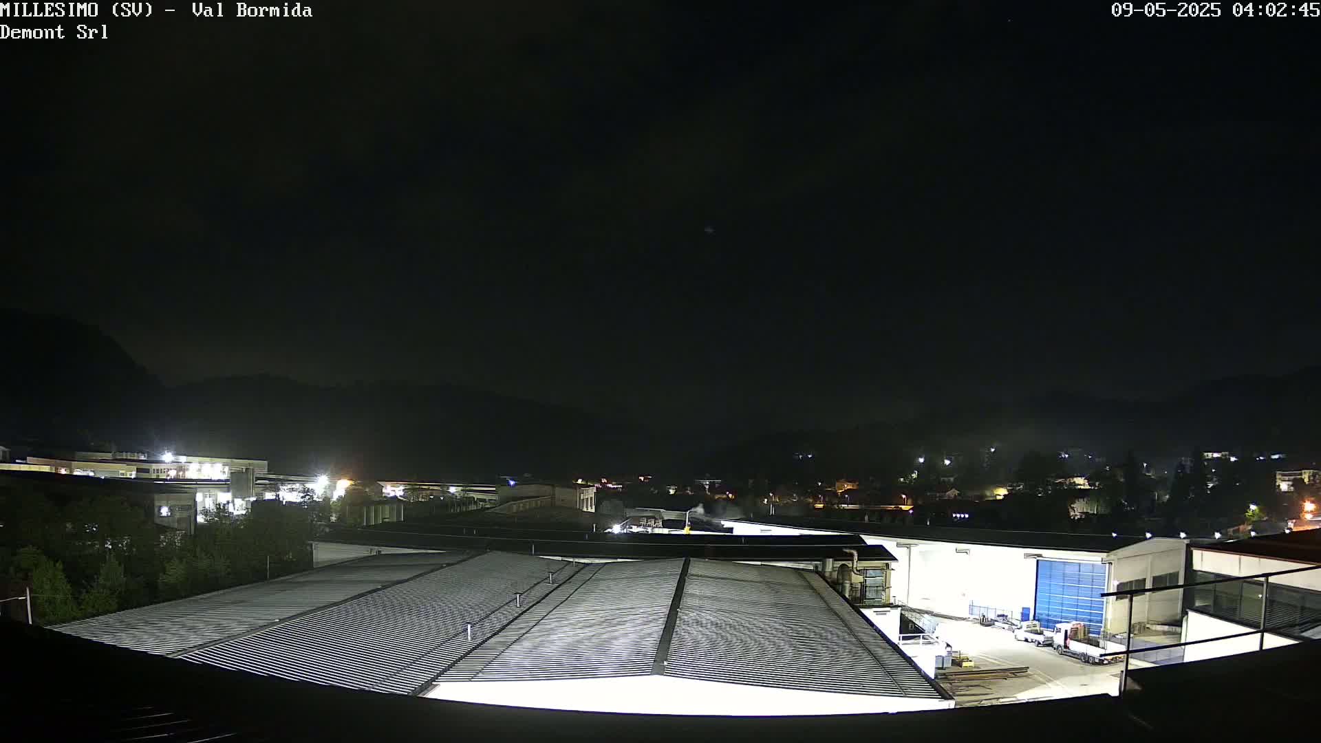 A nighttime, high-angle view shows a large industrial area with several buildings, including a factory with a corrugated metal roof, under a dark, clear sky.