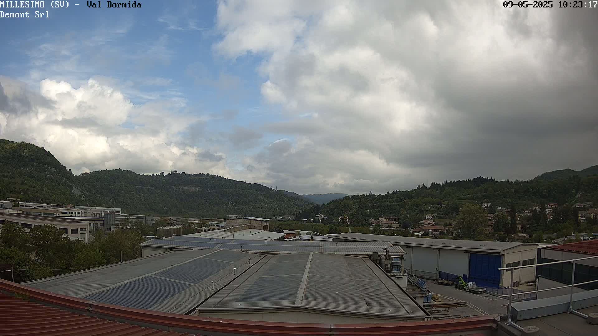 A cloudy day reveals a view of industrial buildings and a town nestled in a valley surrounded by green hills.
