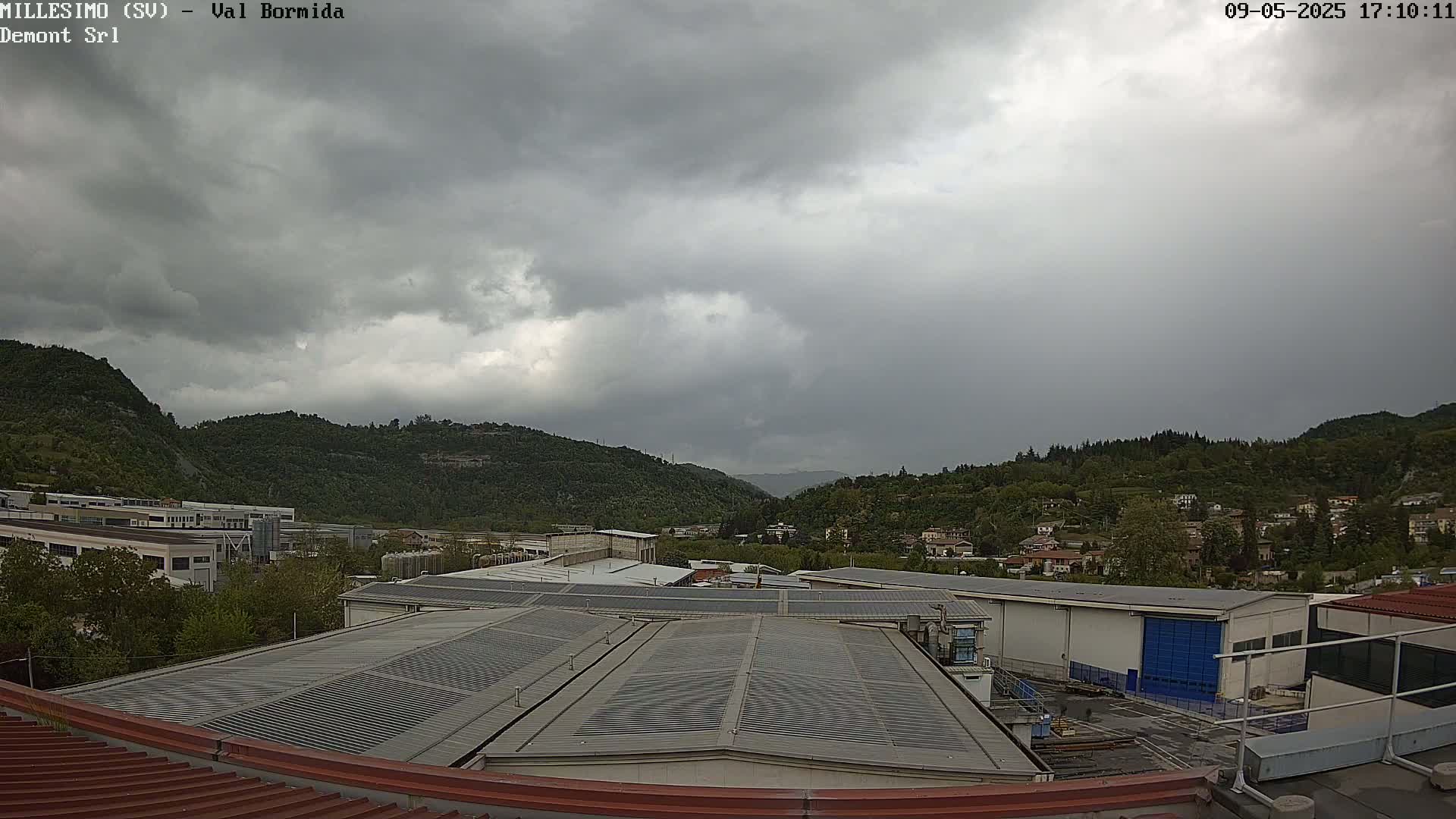 Overcast skies loom over a valley of green hills and industrial buildings, viewed from a rooftop.