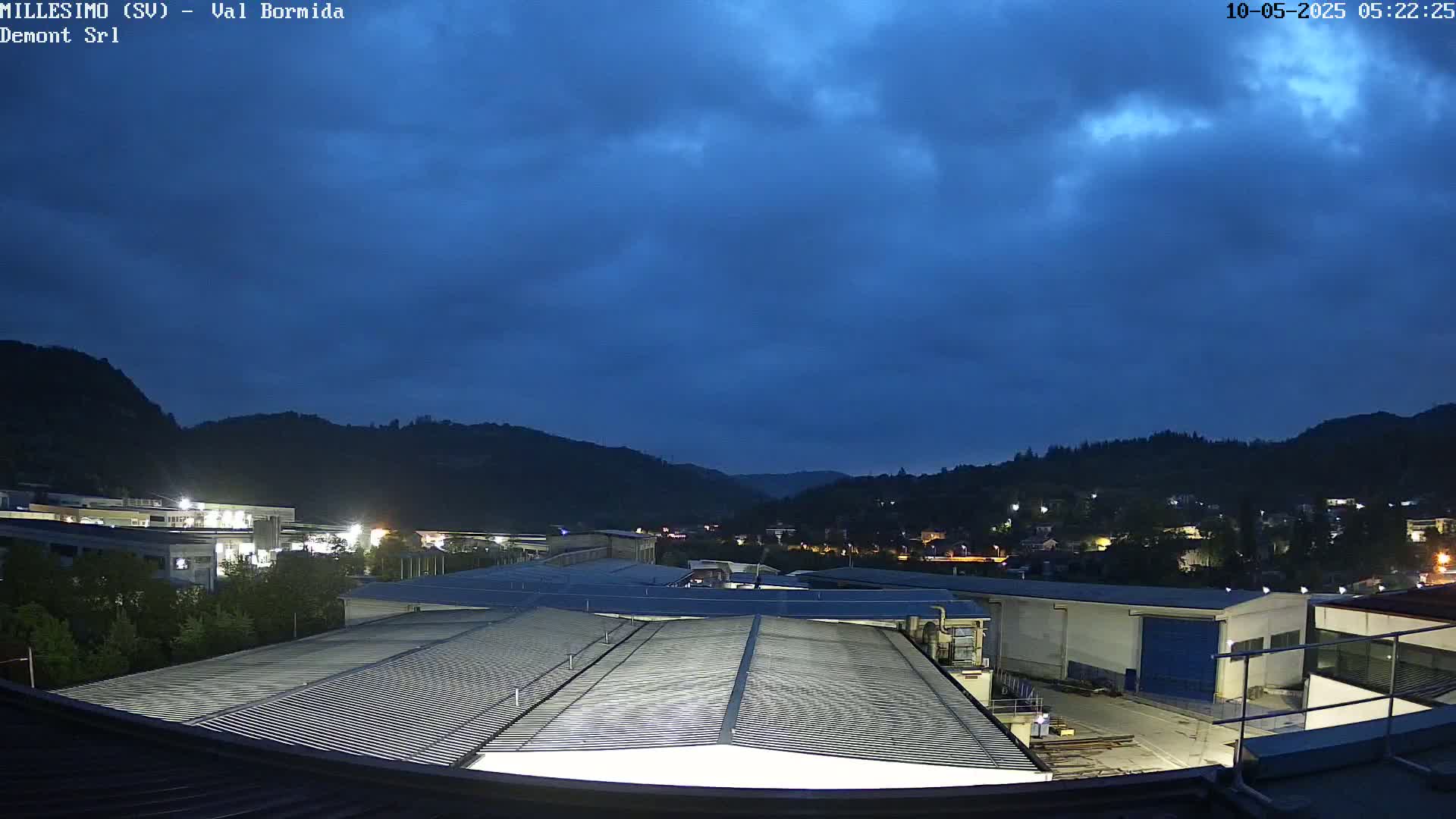 Under a dark blue, cloudy sky, an industrial area with several buildings is nestled in a valley surrounded by hills.