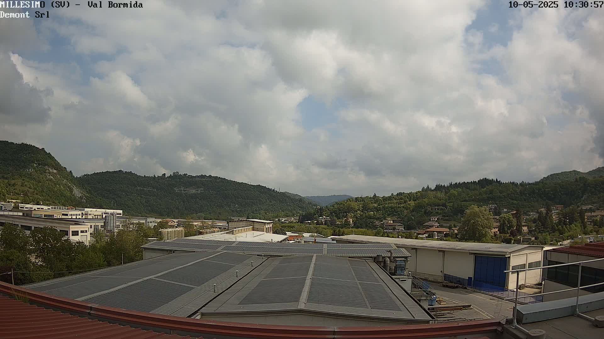 A partly cloudy sky overlooks a valley of green hills and industrial buildings, viewed from a rooftop with a gray metal roof.