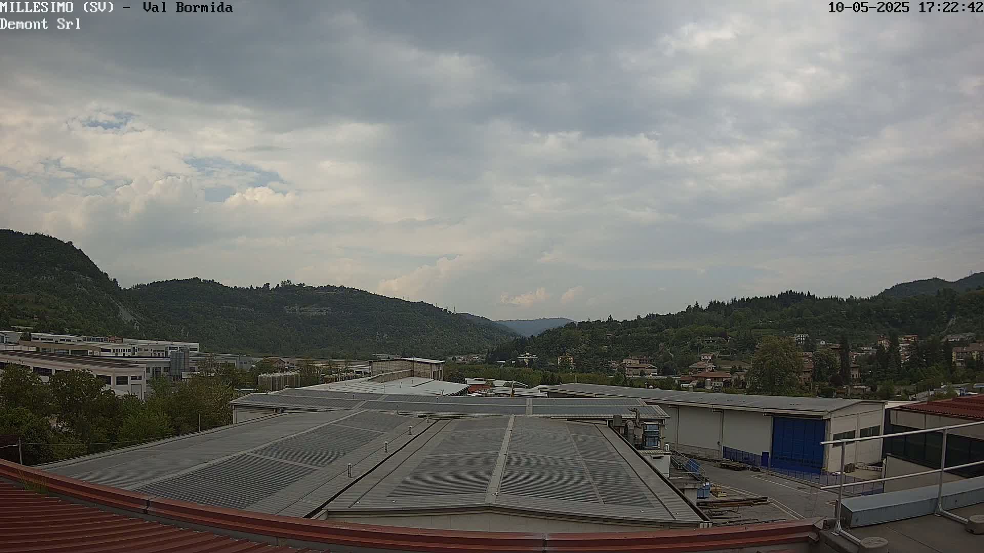 A mostly cloudy sky overlooks an industrial area nestled in a valley between green hills.