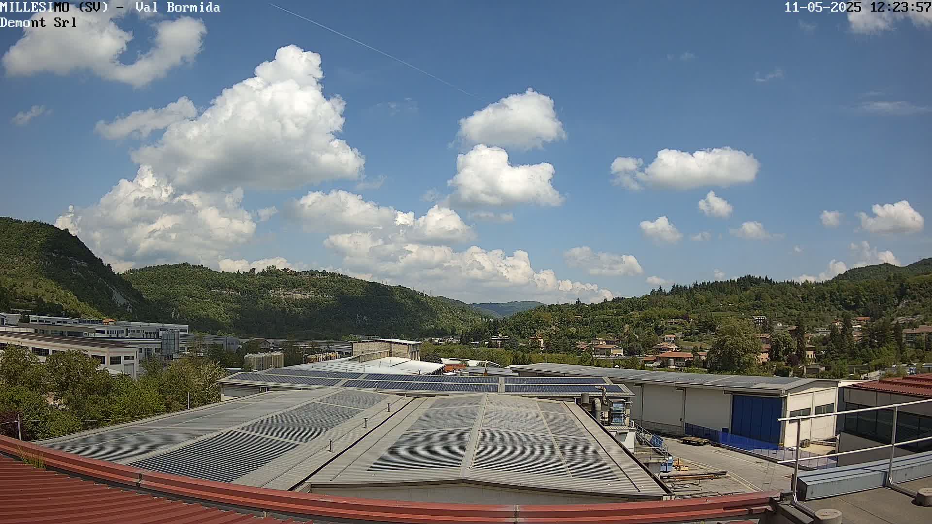 A partly cloudy sky overlooks industrial buildings and a residential area nestled in a valley surrounded by green hills.