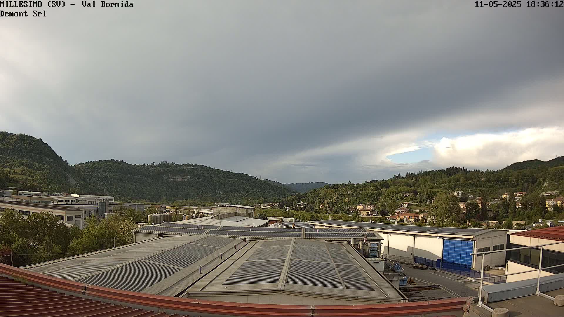 A view from a rooftop shows industrial buildings and a residential area nestled in a valley surrounded by green hills under an overcast sky.