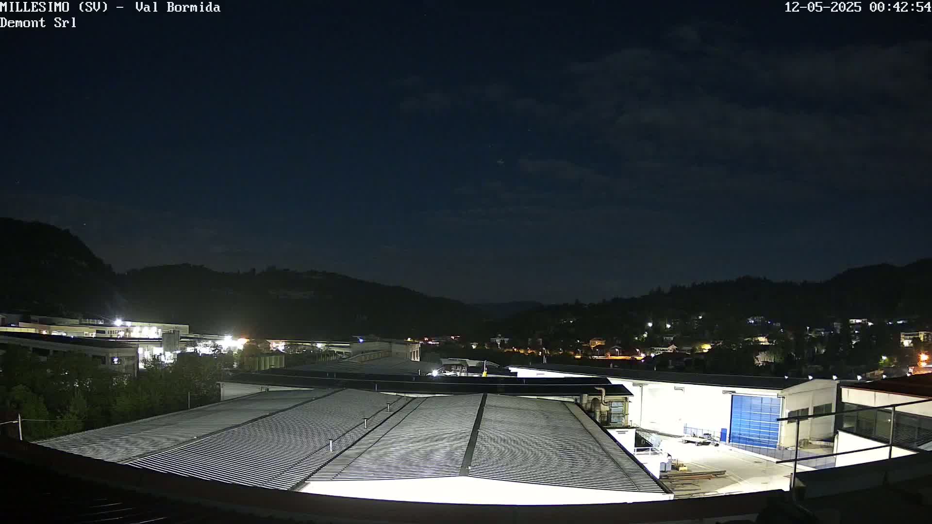 A nighttime view of an industrial area nestled in a valley under a partly cloudy sky.
