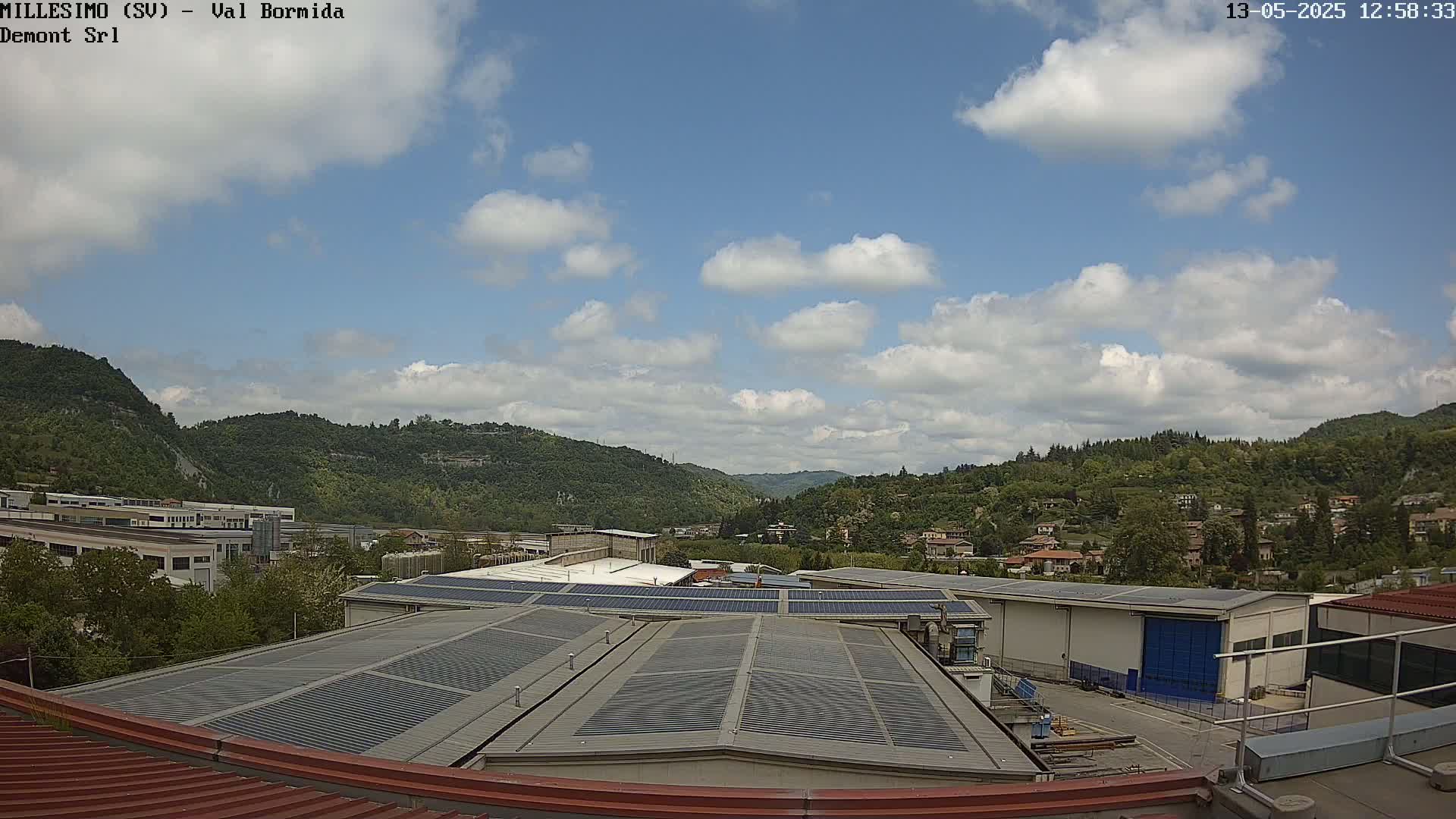 A partly cloudy sky overlooks a valley of green hills dotted with buildings and industrial complexes, with solar panels prominent on a nearby rooftop.