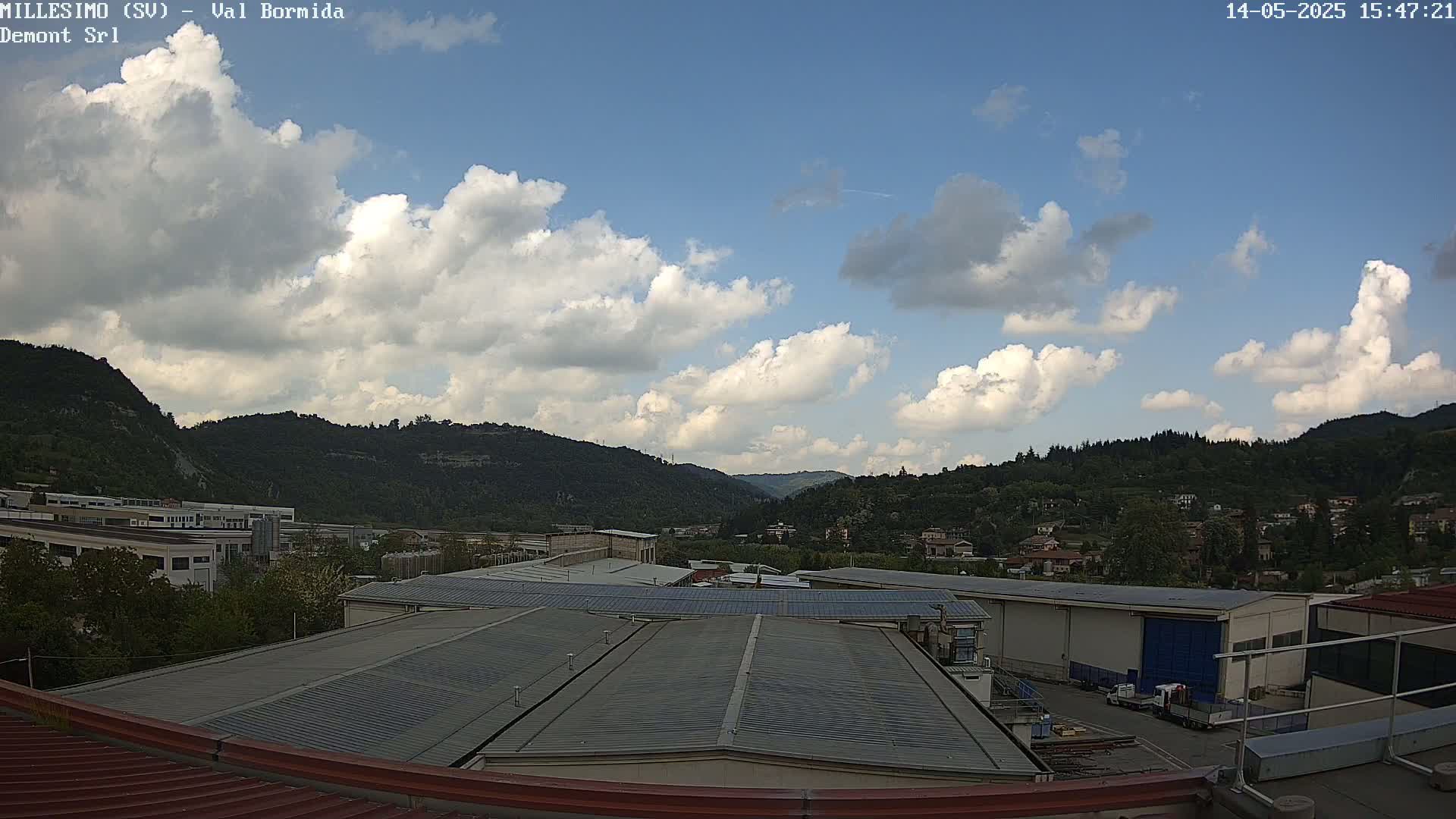 A partly cloudy sky overlooks a valley of green hills and industrial buildings with flat roofs.