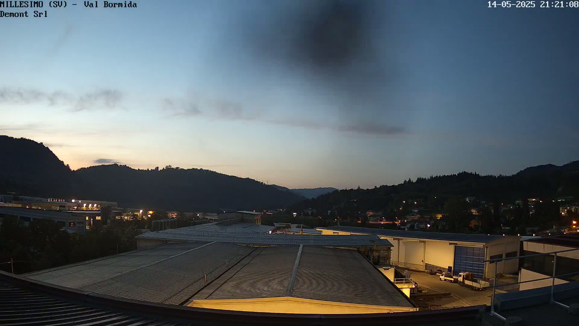 A twilight view shows industrial buildings and a town nestled in a valley between dark hills under a mostly clear, dusky sky.
