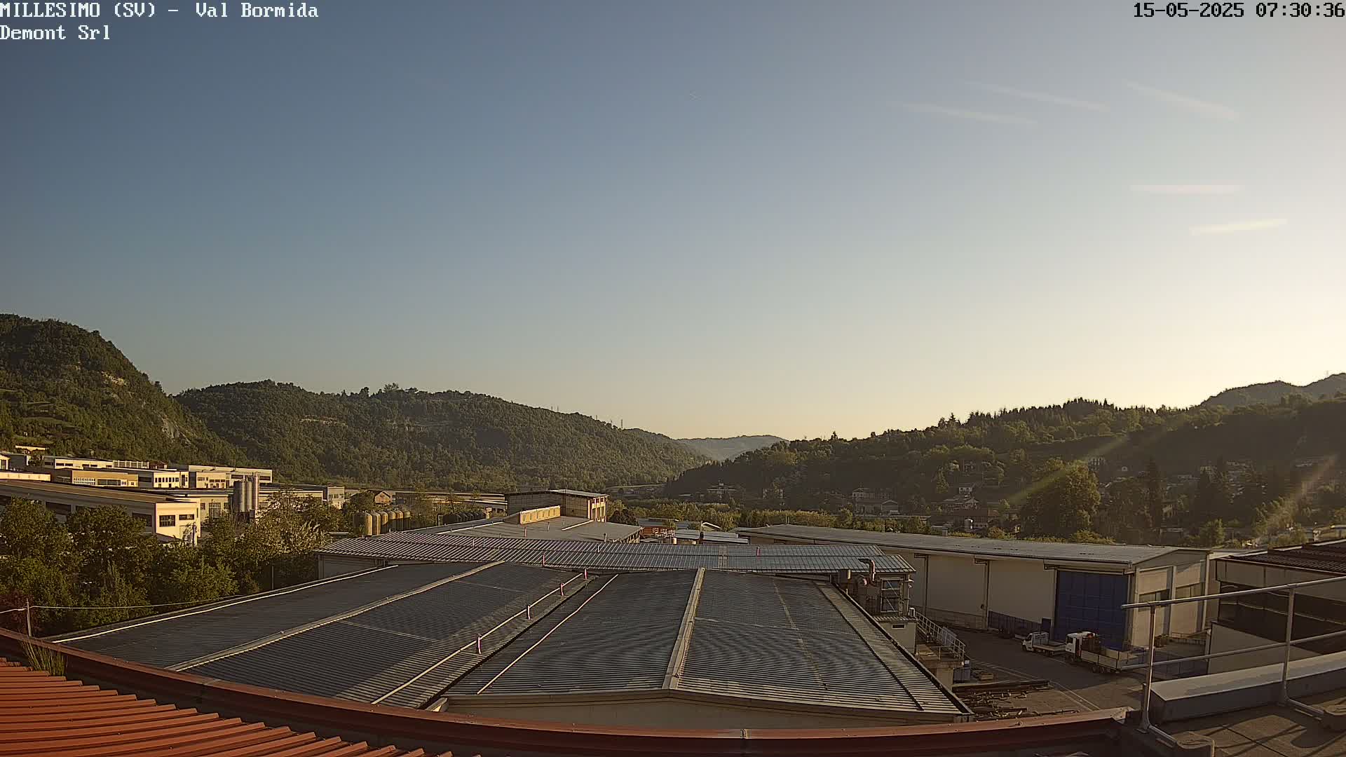 A clear, sunny view of industrial buildings and forested hills in the distance.
