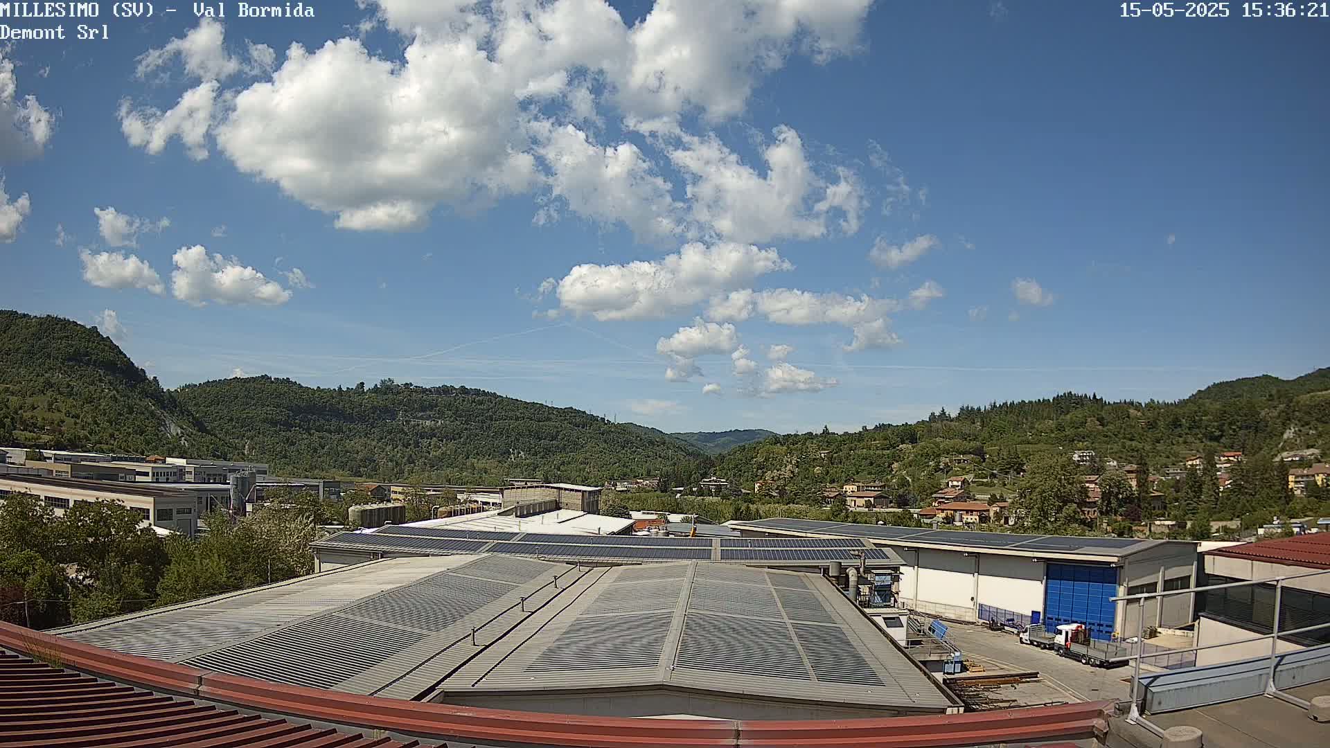A partly cloudy sky overlooks a valley of green hills and industrial buildings with solar panels on their roofs.