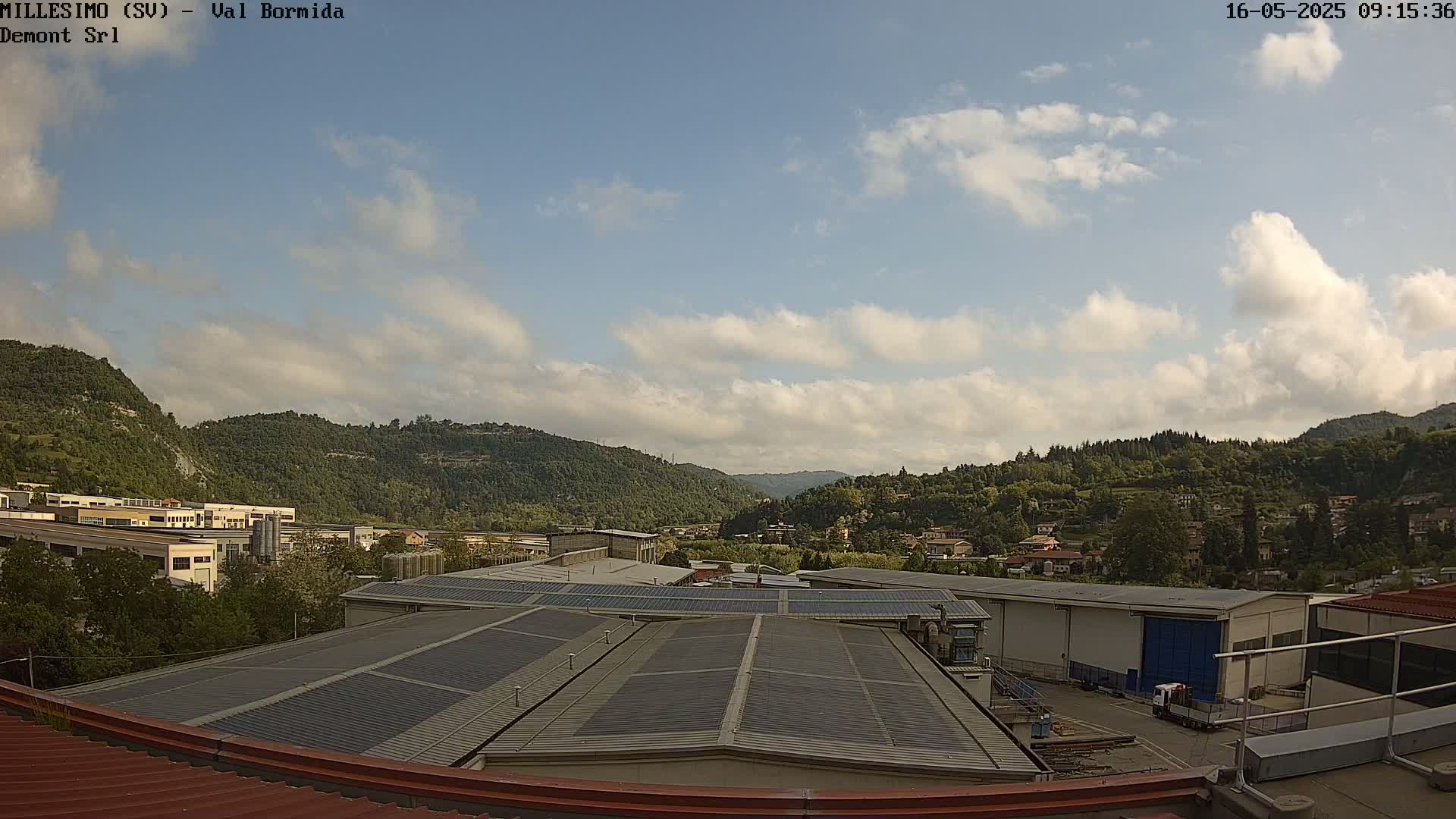 A partly cloudy sky overlooks a valley of green hills and industrial buildings, with solar panels on a rooftop in the foreground.